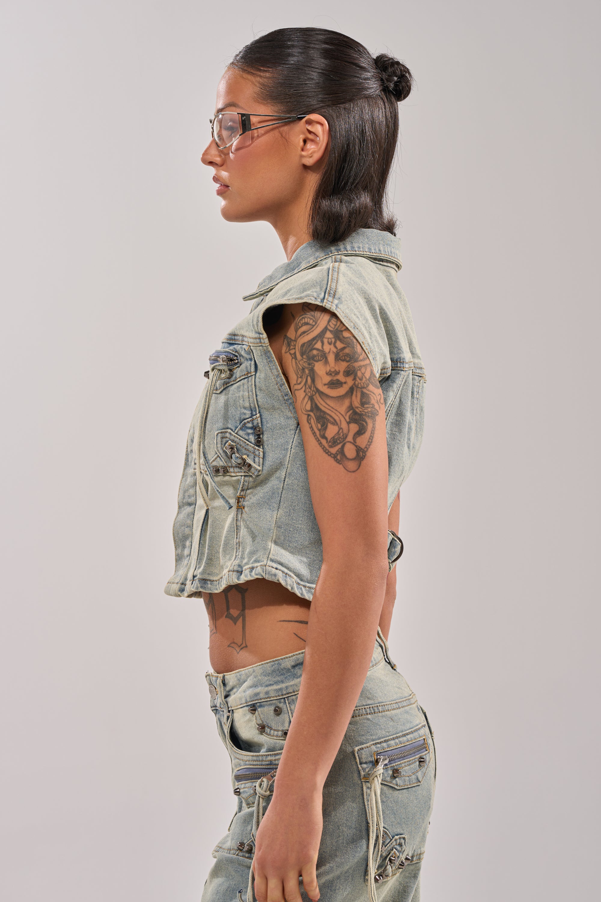 A woman with straight dark hair tied back, wearing clear glasses and the COMEBACK ERA DENIM SLEEVELESS TOP over matching denim pants, stands in profile revealing a large upper arm tattoo against a plain background, exuding effortless street style. 2