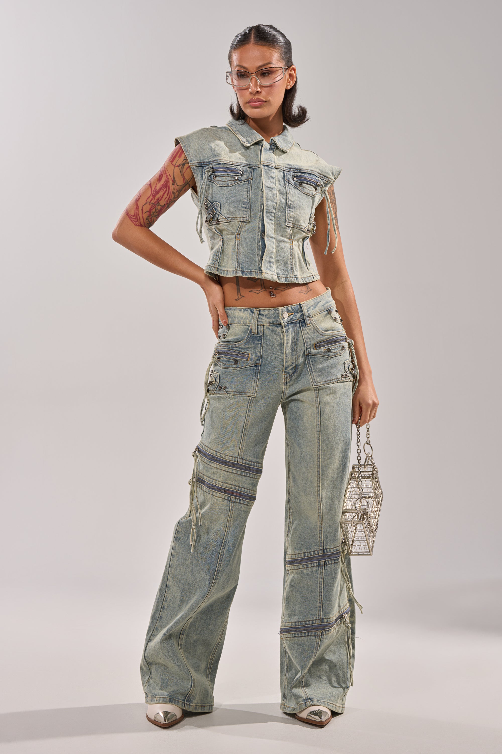 A woman displays street style in the COMEBACK ERA DENIM SLEEVELESS TOP and matching wide-leg pants with zippers and pockets, accessorized with clear glasses, white shoes, and a geometric metallic handbag. 4