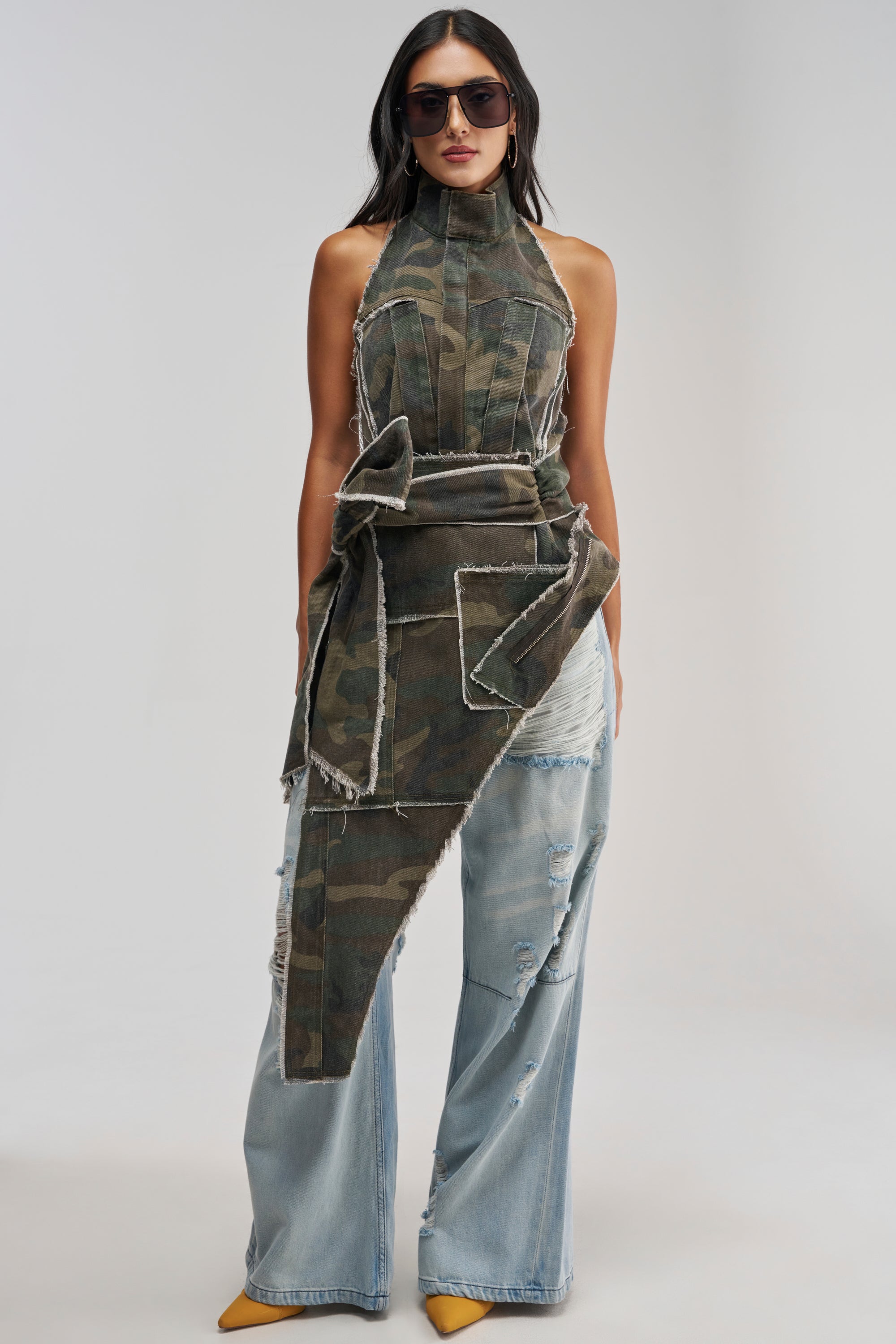 Wearing the BIG STEPPER CAMO TOP—a halter blouse with asymmetrical layers and frayed edges—styled as a festival outfit with sunglasses, ripped wide-leg jeans, and yellow pointed shoes, standing against a plain background.