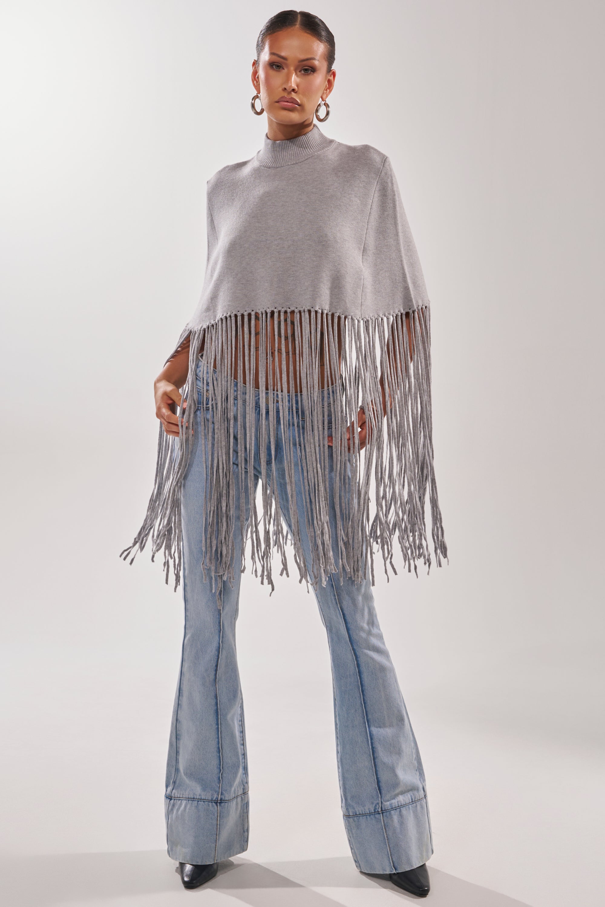A woman models the FRINGE FOR DAYS SWEATER in light gray, paired with light blue flared jeans and black pointed-toe boots, posing confidently against a plain light background.