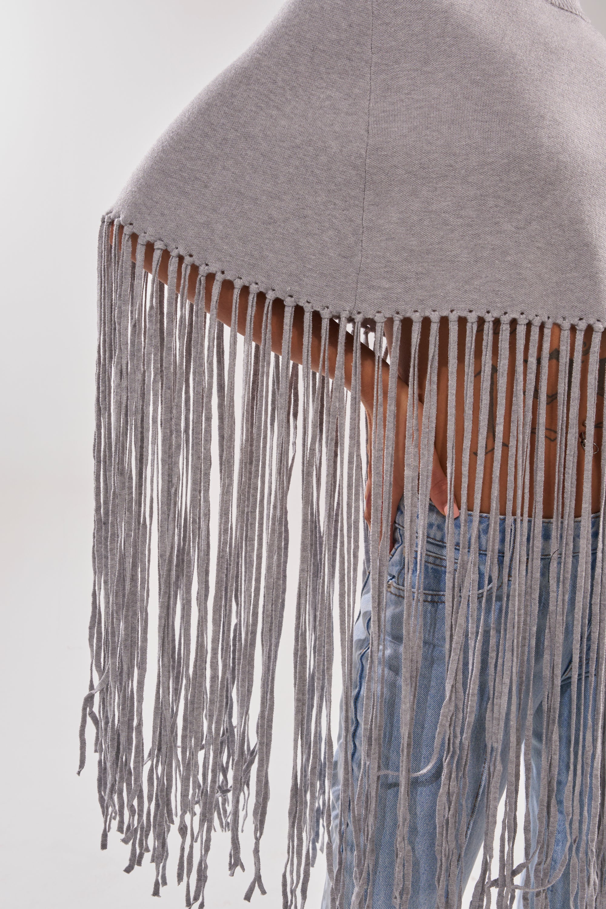 Someone is wearing the FRINGE FOR DAYS SWEATER, a light gray poncho with long, thick fringe that hangs below the waist, styled over light blue jeans against a plain, light background.