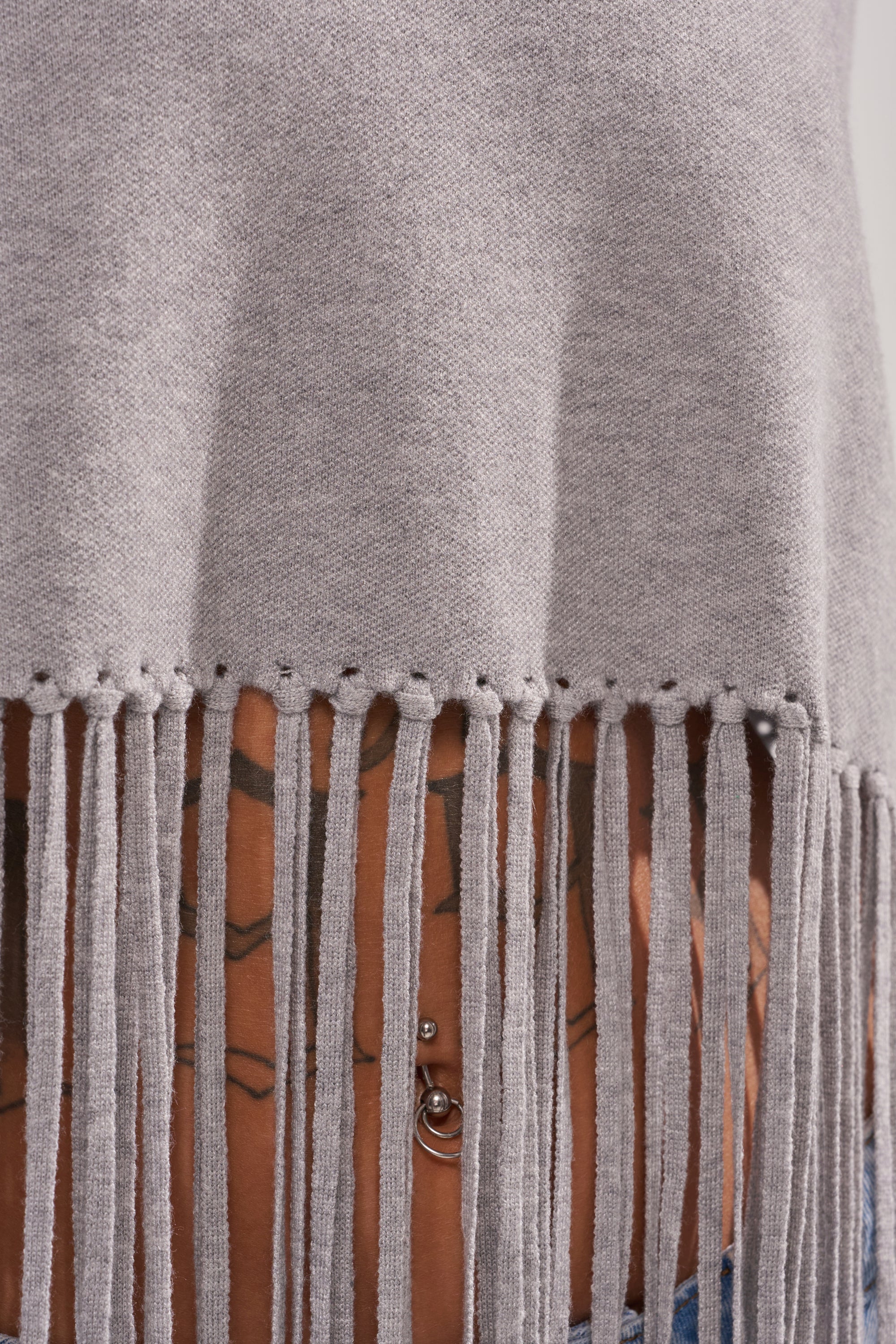 A close-up of a person wearing the FRINGE FOR DAYS SWEATER, a gray knit with long, draping fringes that reveal tattoos and a navel piercing, emphasizing the unique style of this statement layering piece.