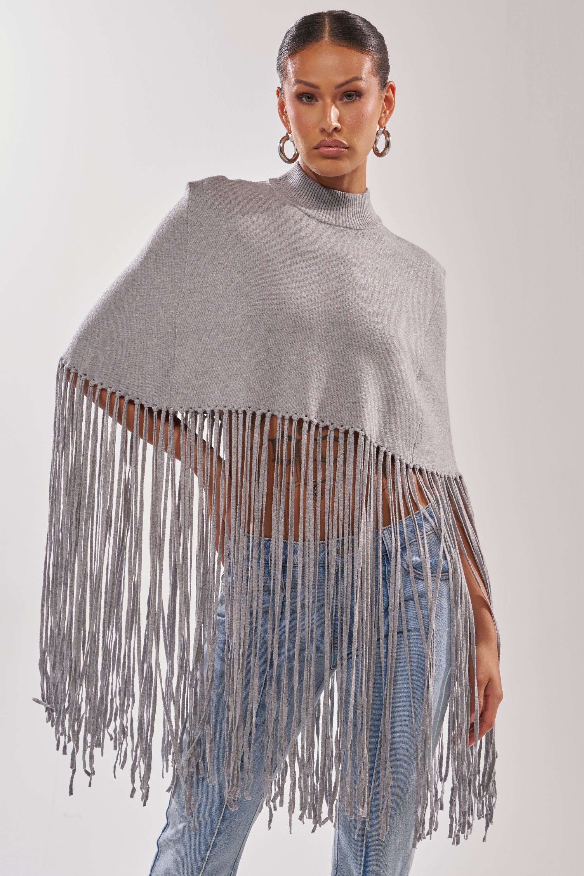 A woman poses confidently in the FRINGE FOR DAYS SWEATER, a gray poncho with long fringe detailing, styled over light blue jeans. With slicked-back hair and hoop earrings, she stands out against a plain light background.
