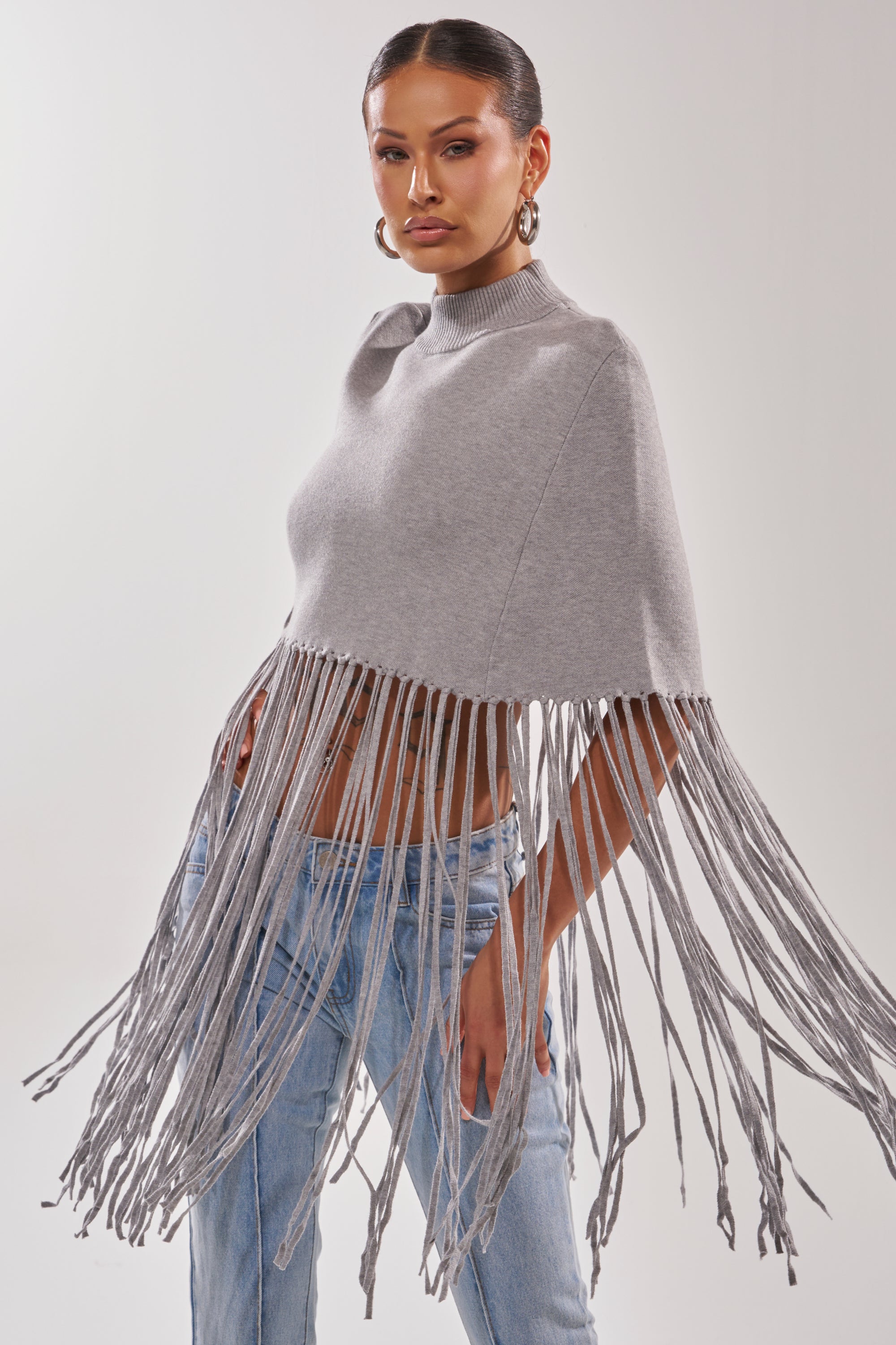 A woman models the FRINGE FOR DAYS SWEATER, a light gray knit poncho with long, thick tassels. Paired with blue jeans and hoop earrings, she stands against a plain backdrop—this chic sweater is the ultimate layering piece.