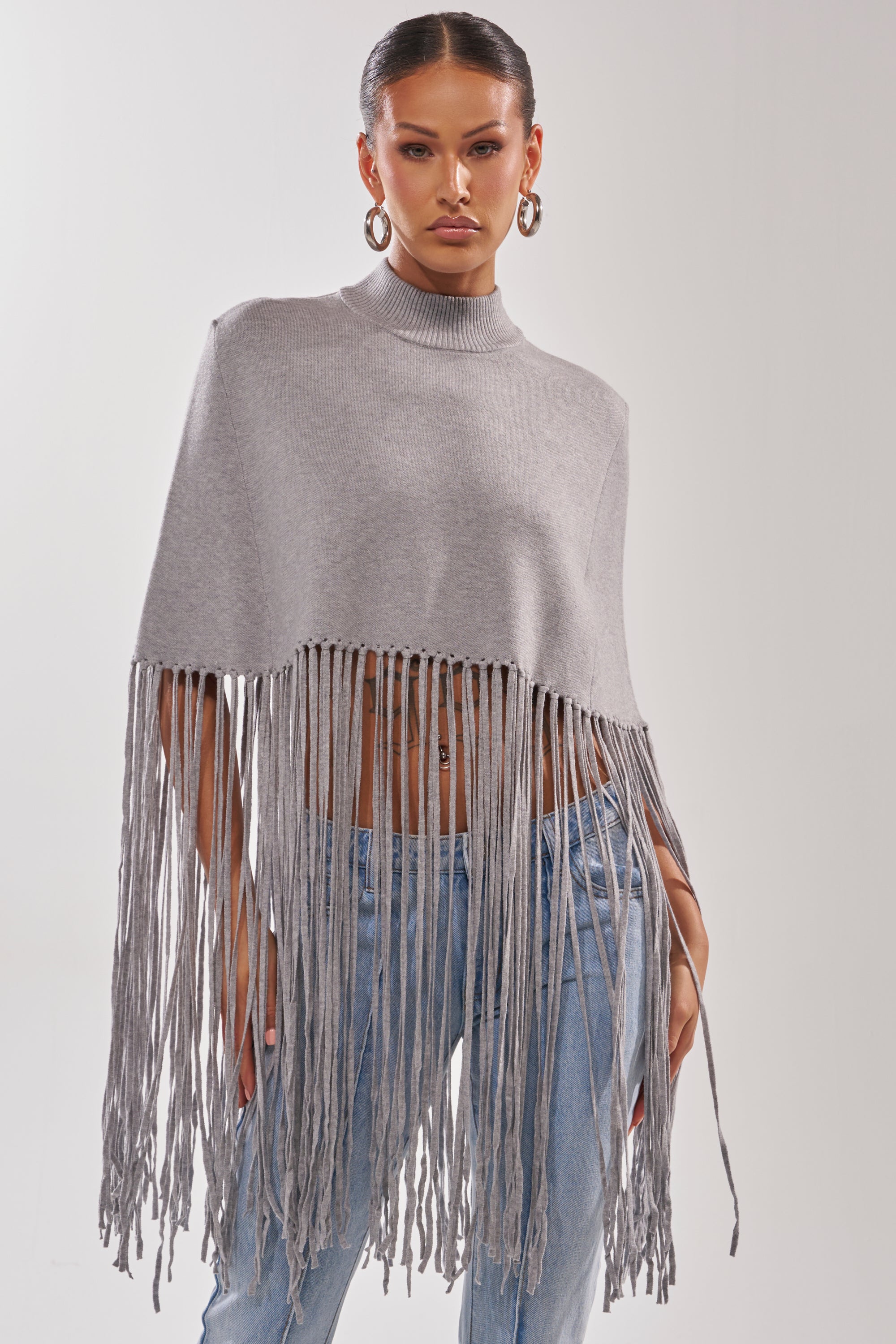 A woman wears hoop earrings, light blue jeans, and the FRINGE FOR DAYS SWEATER—a gray poncho with long fringe details—standing against a plain light background.
