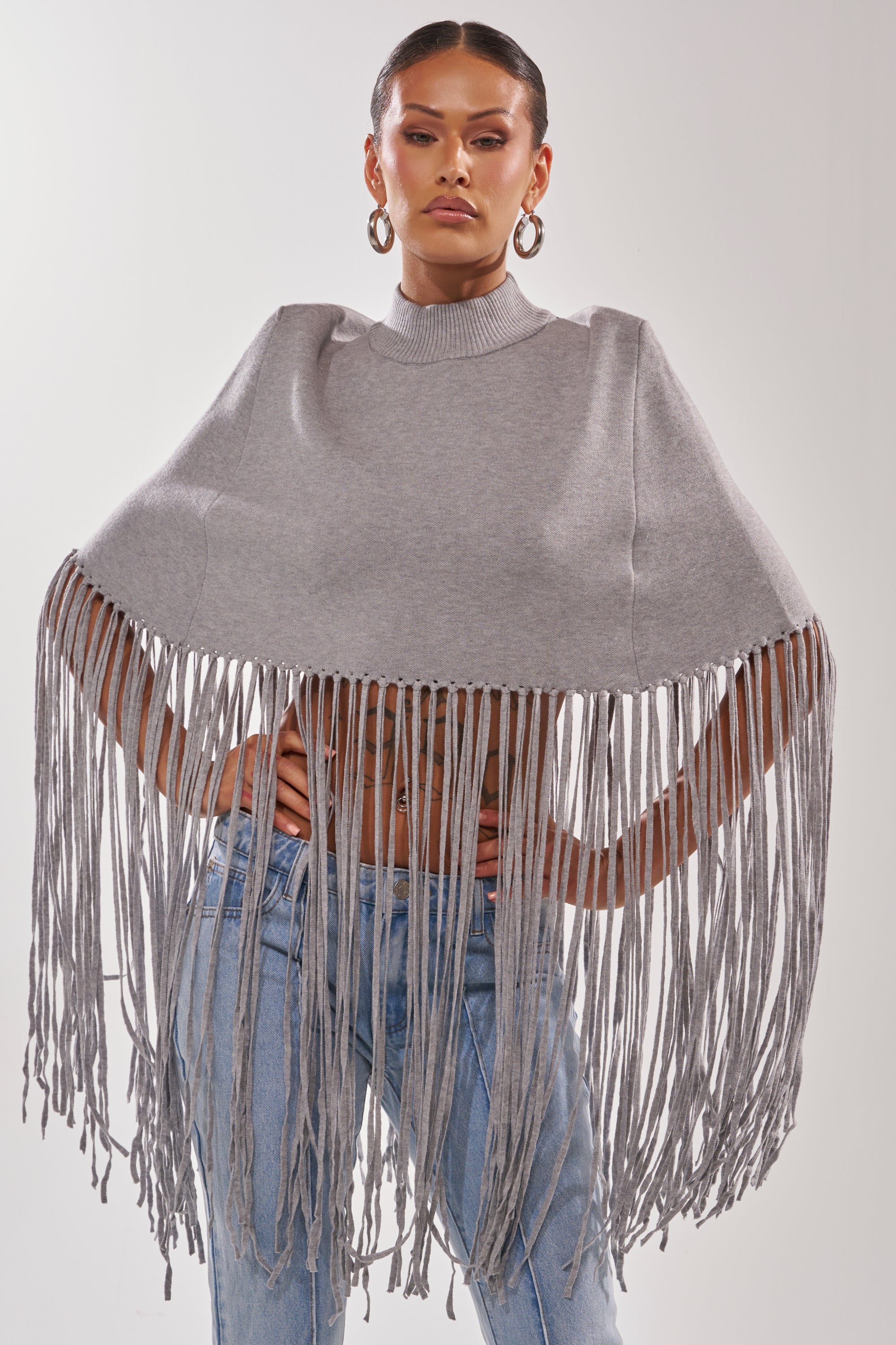 Wearing the FRINGE FOR DAYS SWEATER in gray with blue jeans, a person stands hands on hips, confidently facing the camera against a plain backdrop.