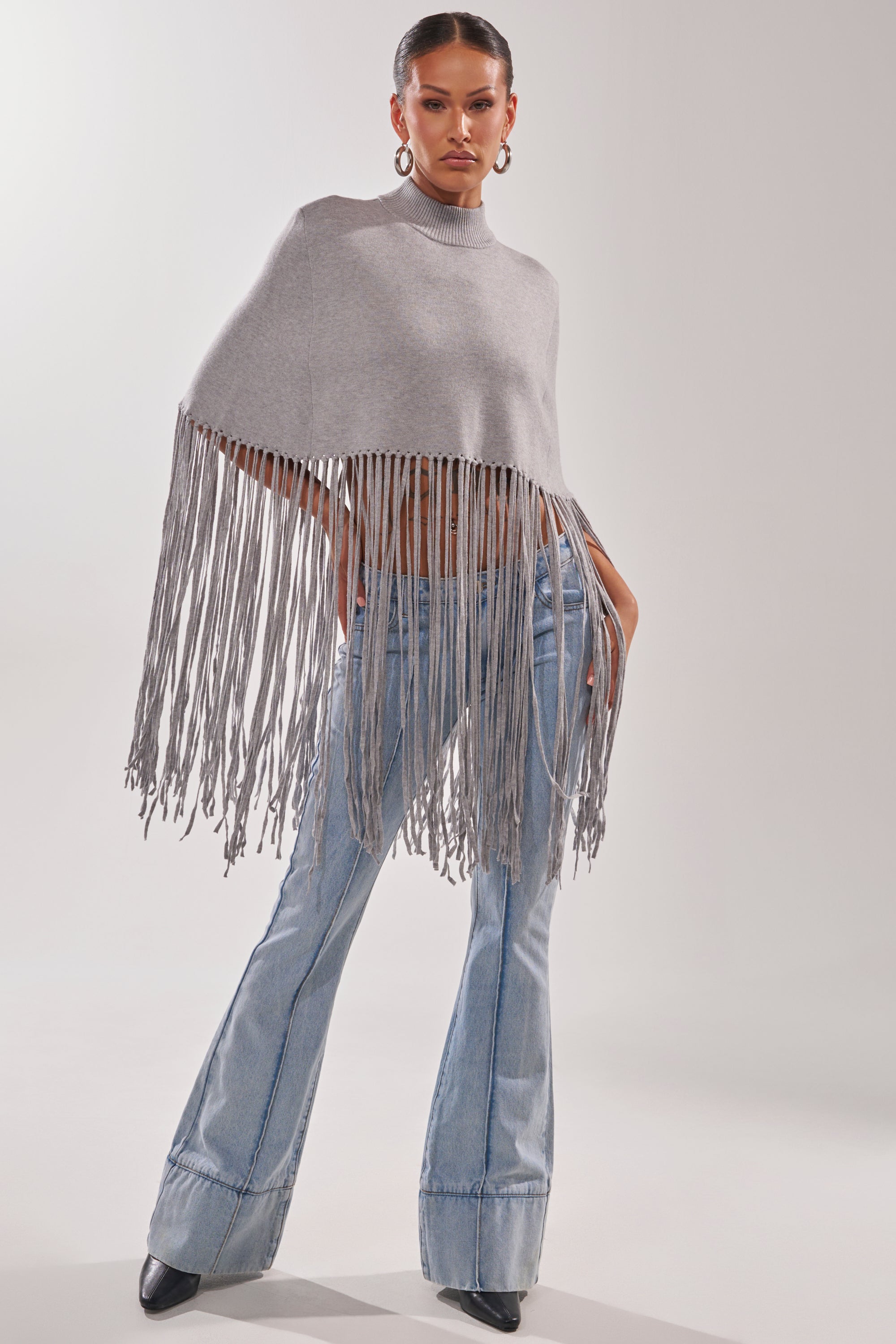 A woman models the FRINGE FOR DAYS SWEATER in light gray with long fringe, paired with light blue flared jeans, hoop earrings, and black pointed-toe shoes as she poses confidently against a plain background.