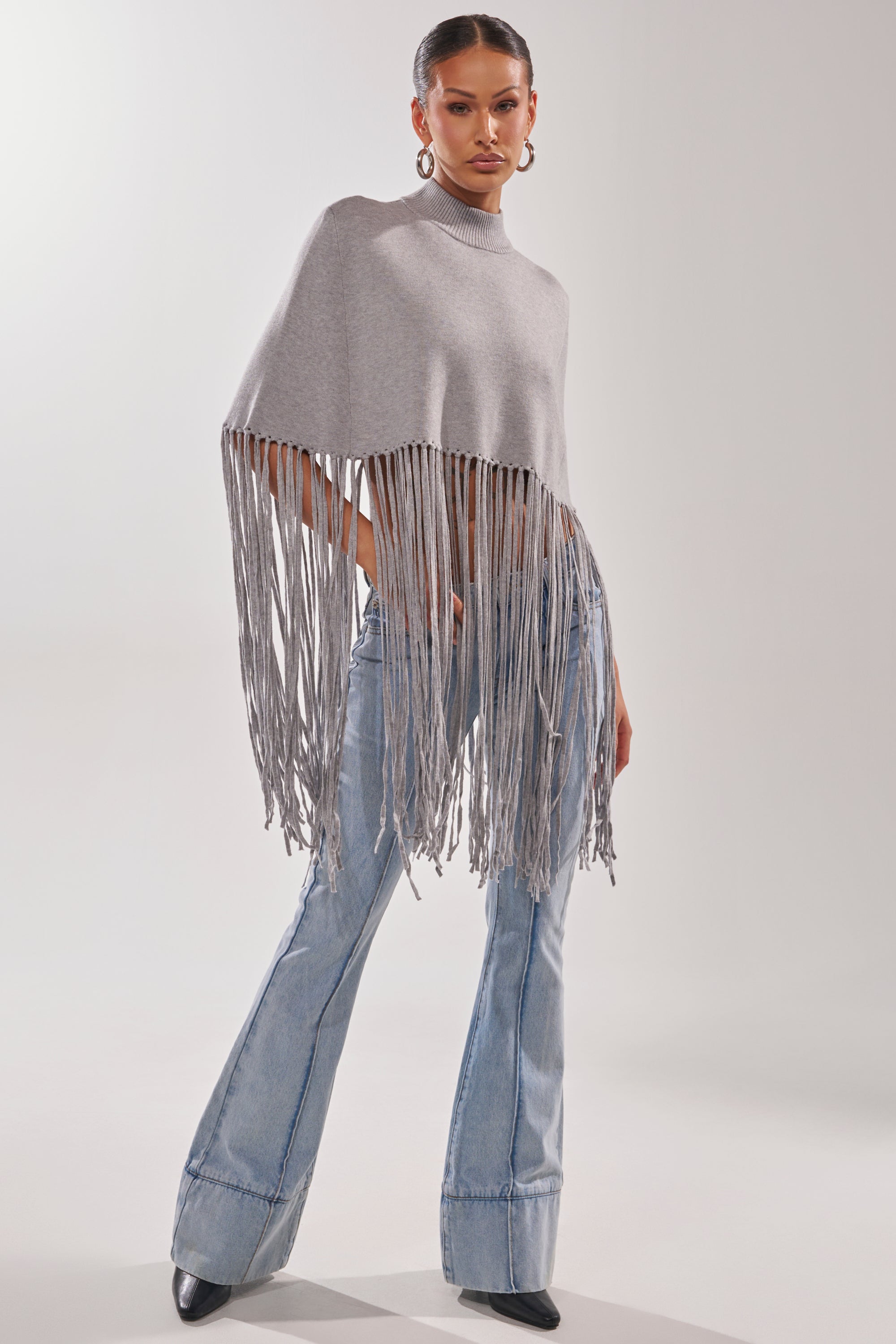 A woman poses confidently against a plain background in the FRINGE FOR DAYS SWEATER, light blue flare jeans, and black heeled boots.