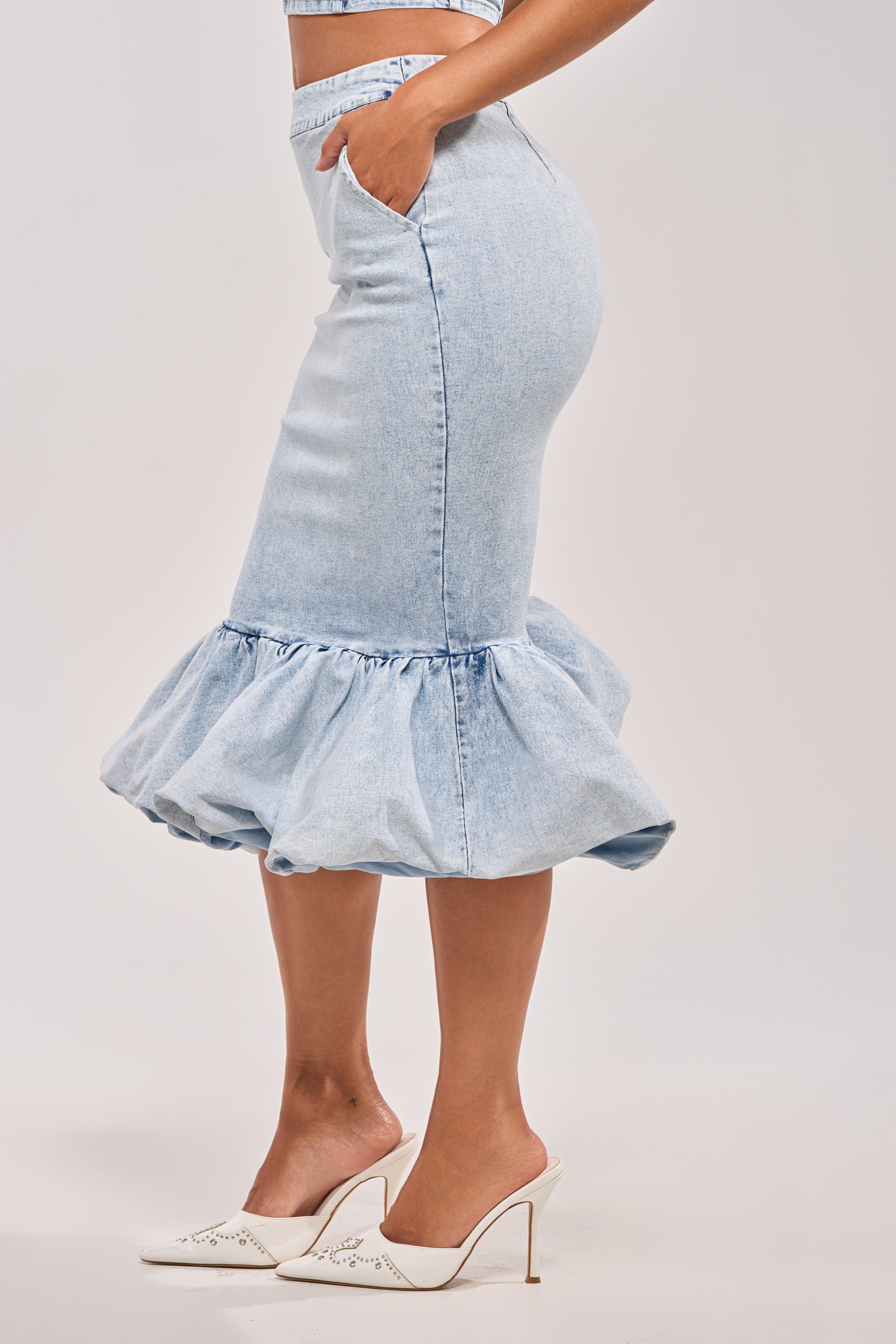 A woman stands in profile wearing the LOVE ME LIKE THAT DENIM MIDI SKIRT, a light blue high-rise midi with a ruffled hem, paired with white high-heeled mules. Her hands are in her pockets against a plain white background. 3