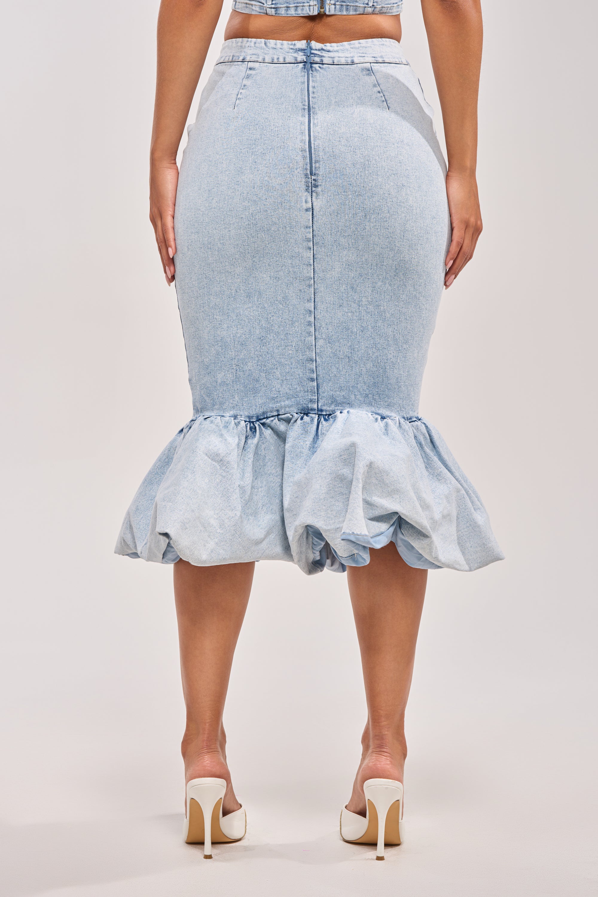 A person is shown from the back against a light background, wearing the LOVE ME LIKE THAT DENIM MIDI SKIRT, which features a light blue wash, high-rise waist, and ruffled hem, paired with white high-heeled shoes. 4