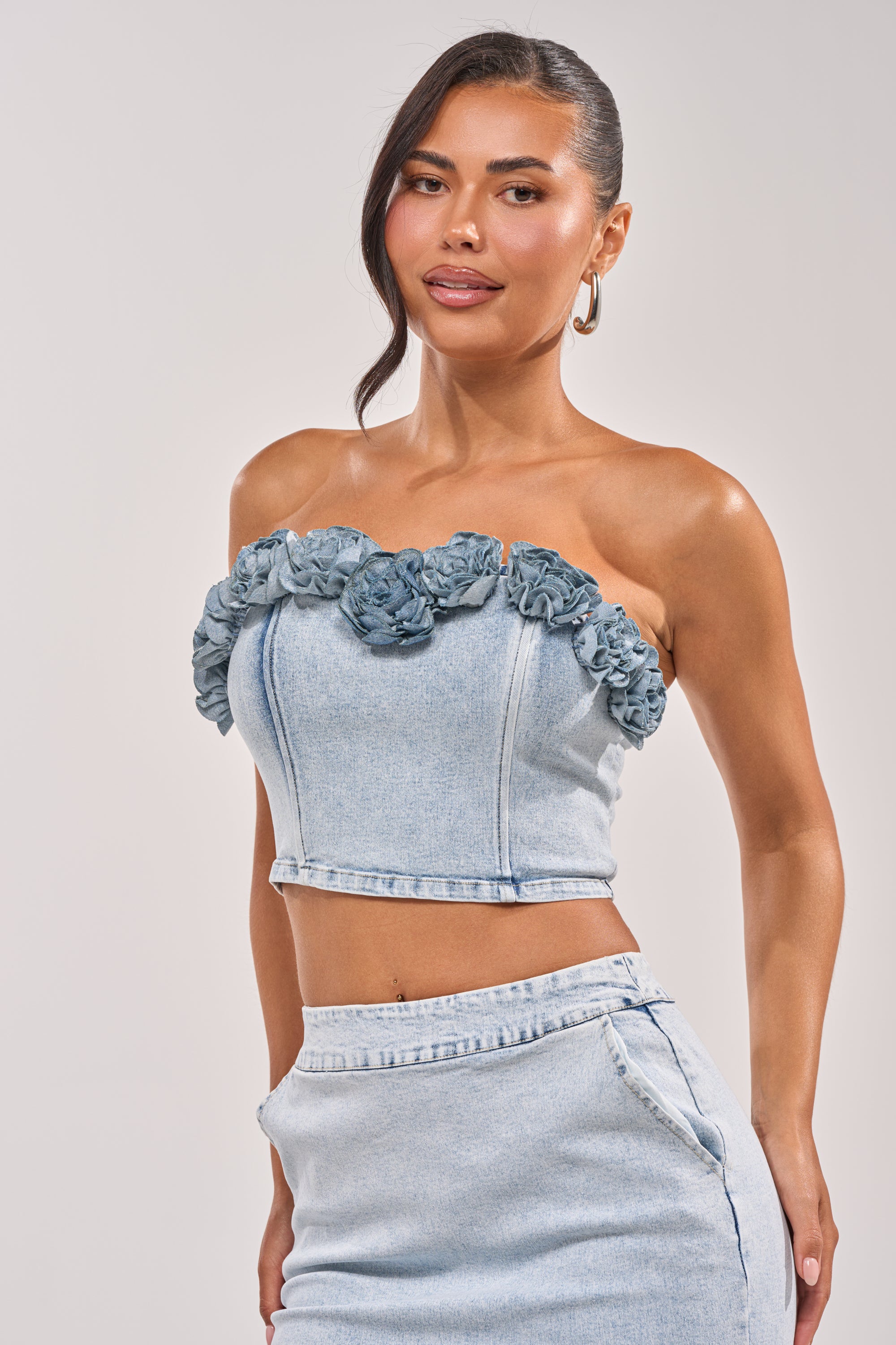 She wears the LOVE ME LIKE THAT FLOWER CORSET, a light denim top with floral neckline detail, paired with a matching high-waisted skirt. Her long dark hair is in a ponytail as she smiles slightly. 0