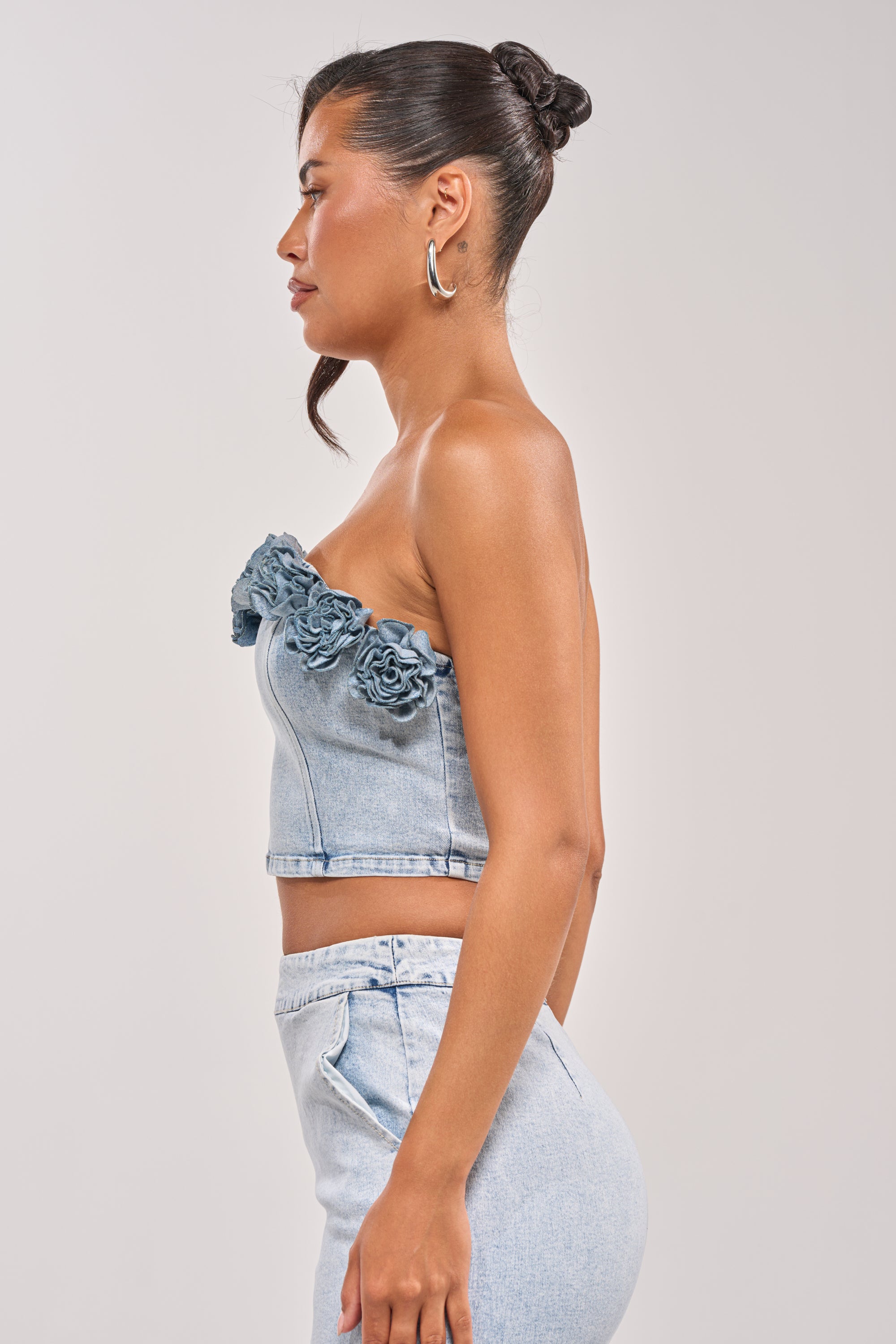 Woman in profile wears the LOVE ME LIKE THAT FLOWER CORSET, a denim corset top with floral details paired with high-waisted denim pants. She completes the look with a sleek bun and hoop earrings against a plain background. 2