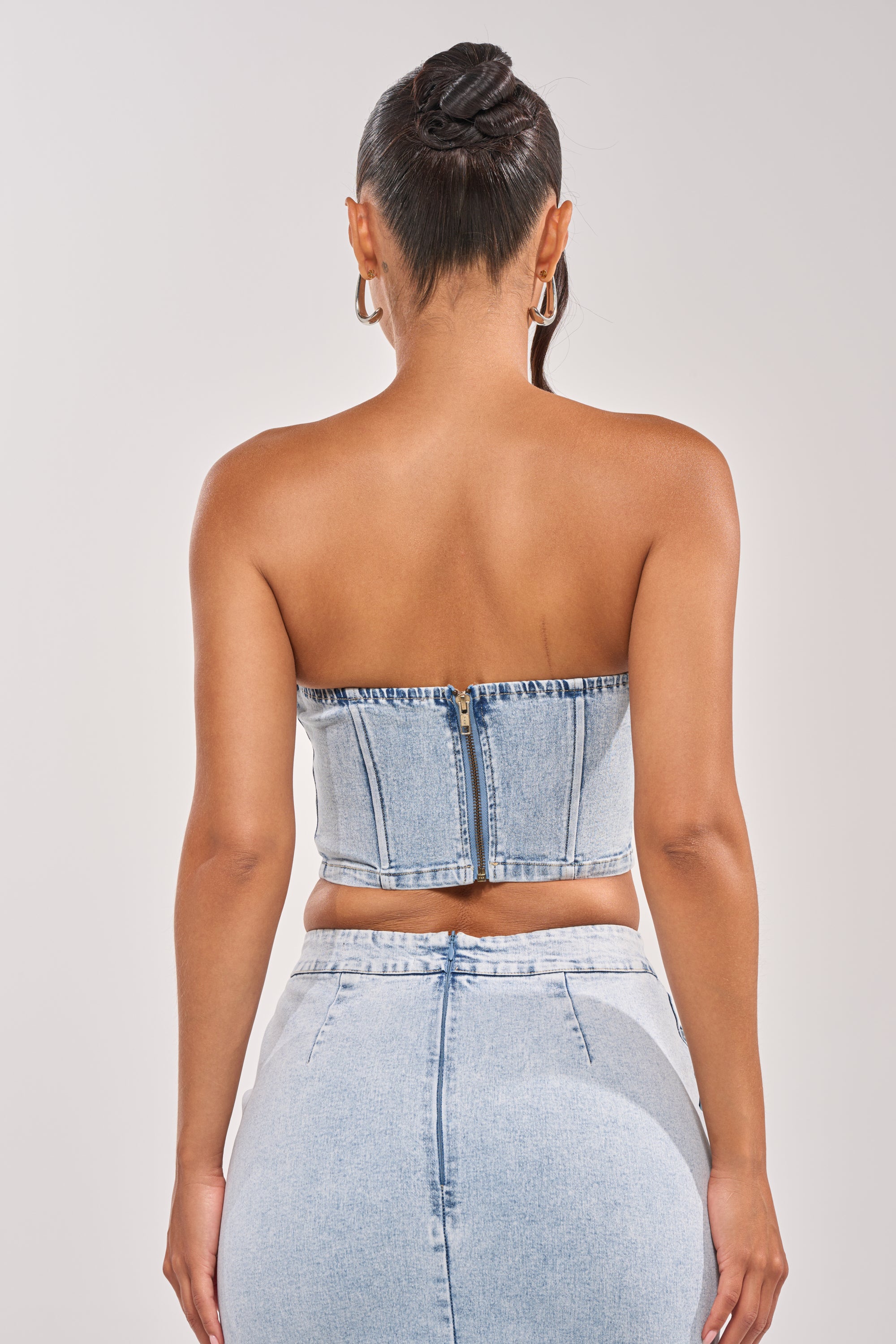A woman faces away from the camera wearing the LOVE ME LIKE THAT FLOWER CORSET, a strapless denim top with a back zipper, paired with a matching high-waisted skirt. Her hair is styled in a sleek bun, and she accessorizes with large hoop earrings. 3