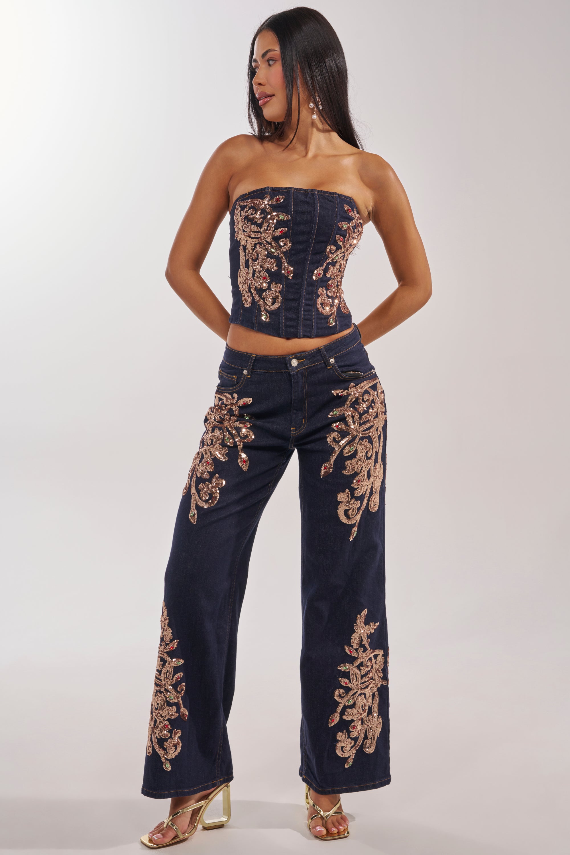 A woman stands with hands on hips, wearing the MALIBU EMBELLISHED WIDE LEG DENIM—a navy blue strapless corset top paired with matching wide-leg pants adorned with intricate gold embroidery—and gold heeled sandals. 0