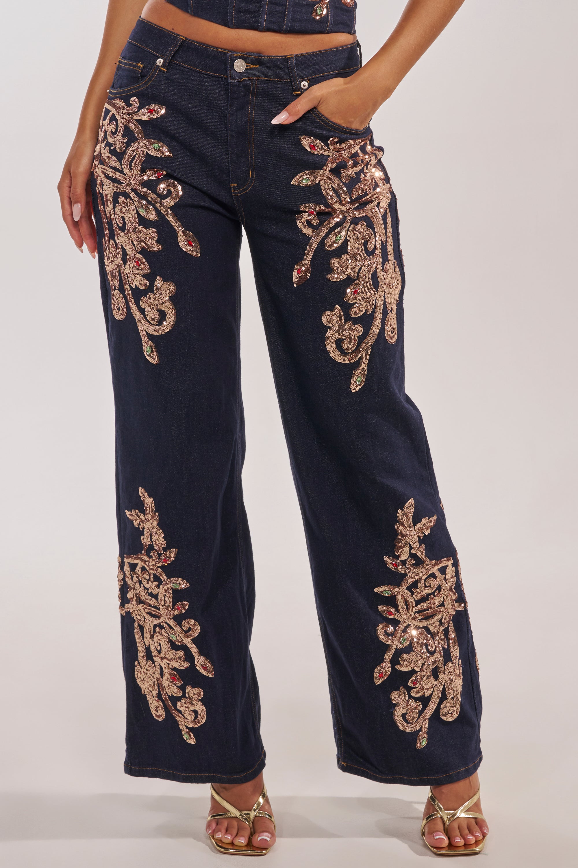 A person wears the MALIBU EMBELLISHED WIDE LEG DENIM with ornate gold sequin embroidery on the pockets and legs, paired with gold strappy heels. The photo is cropped at the torso and head. 1