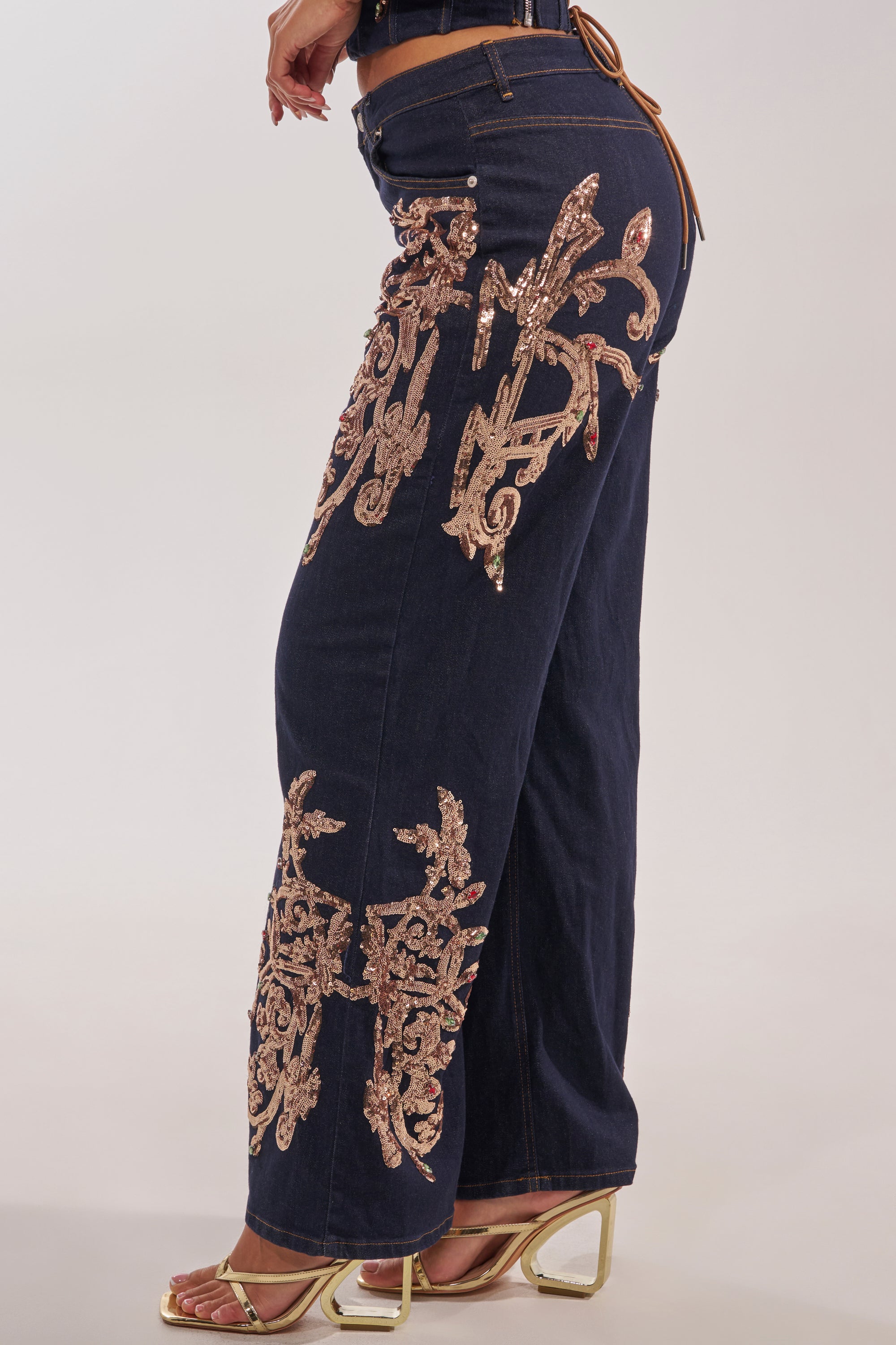 A person wearing MALIBU EMBELLISHED WIDE LEG DENIM with intricate gold sequin details and metallic gold high-heeled sandals, shown from the side against a plain background. 3