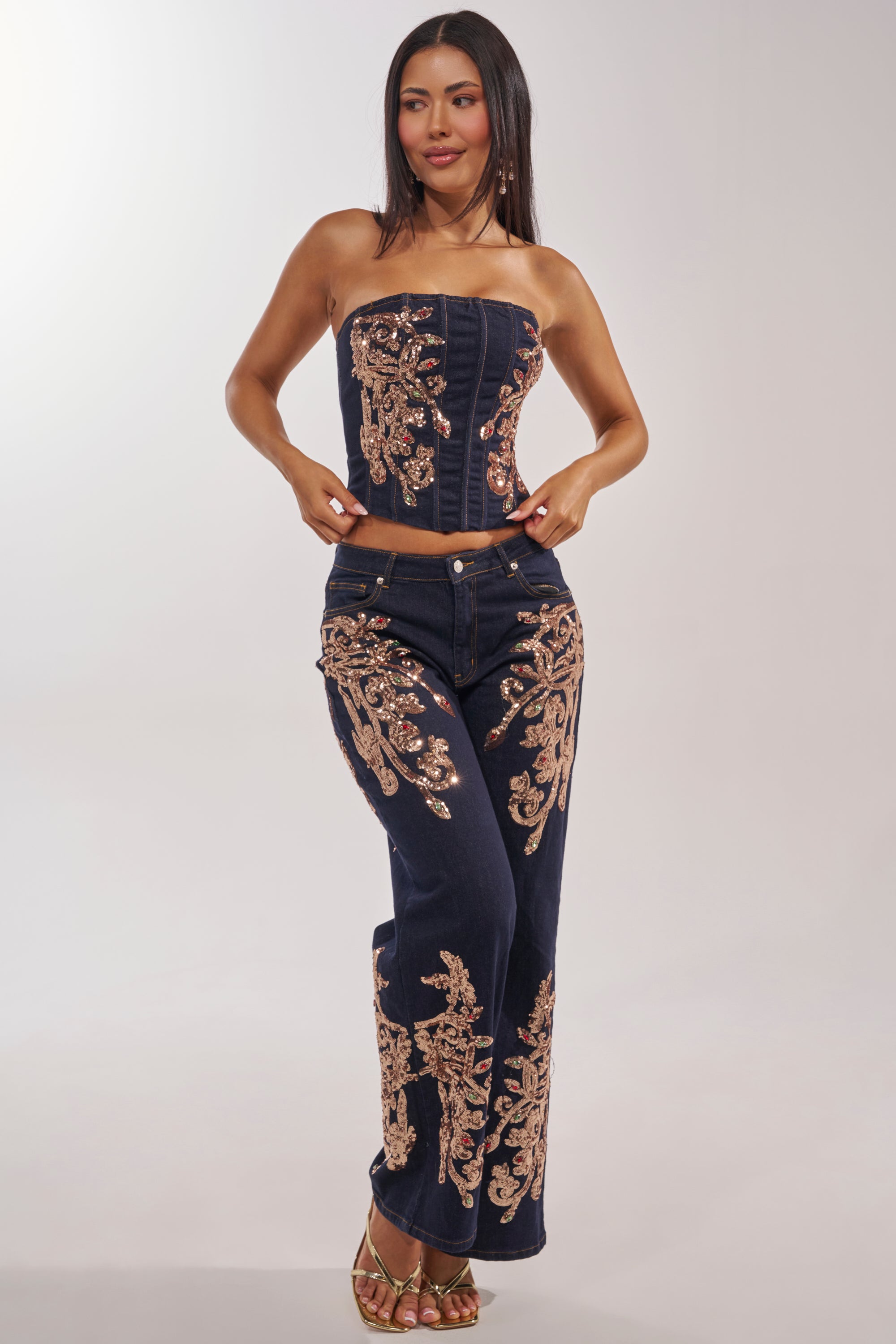 A woman poses with one hand on her hip, smiling slightly, wearing the MALIBU EMBELLISHED WIDE LEG DENIM—a navy blue wide-leg jean adorned with gold sequin embroidery. She pairs it with a matching strapless corset top and has long straight hair. 5
