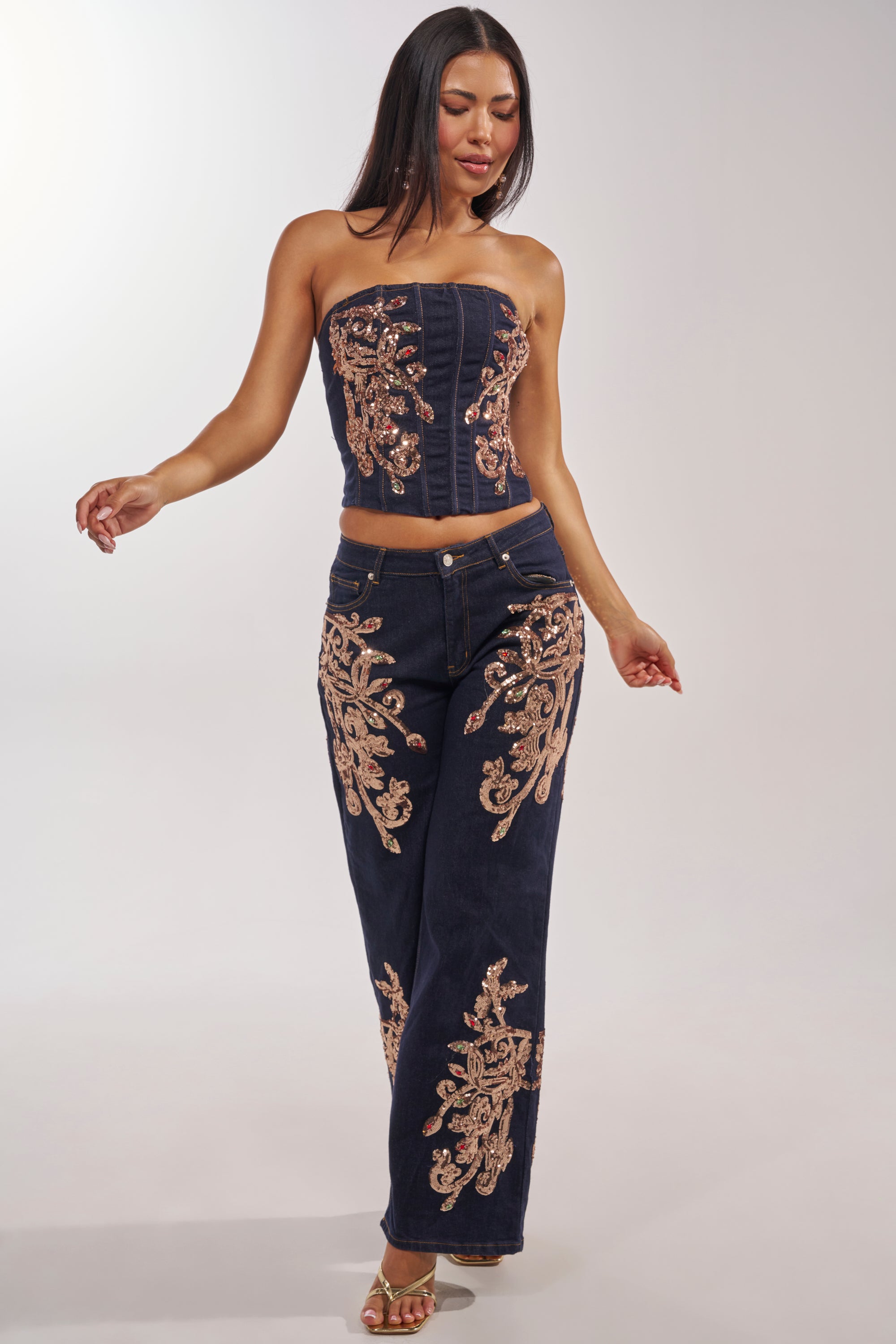 A woman models the MALIBU EMBELLISHED WIDE LEG DENIM pants and a matching strapless corset top, both in navy blue with gold floral embroidery. She completes the look with long dark hair and gold heels. 6
