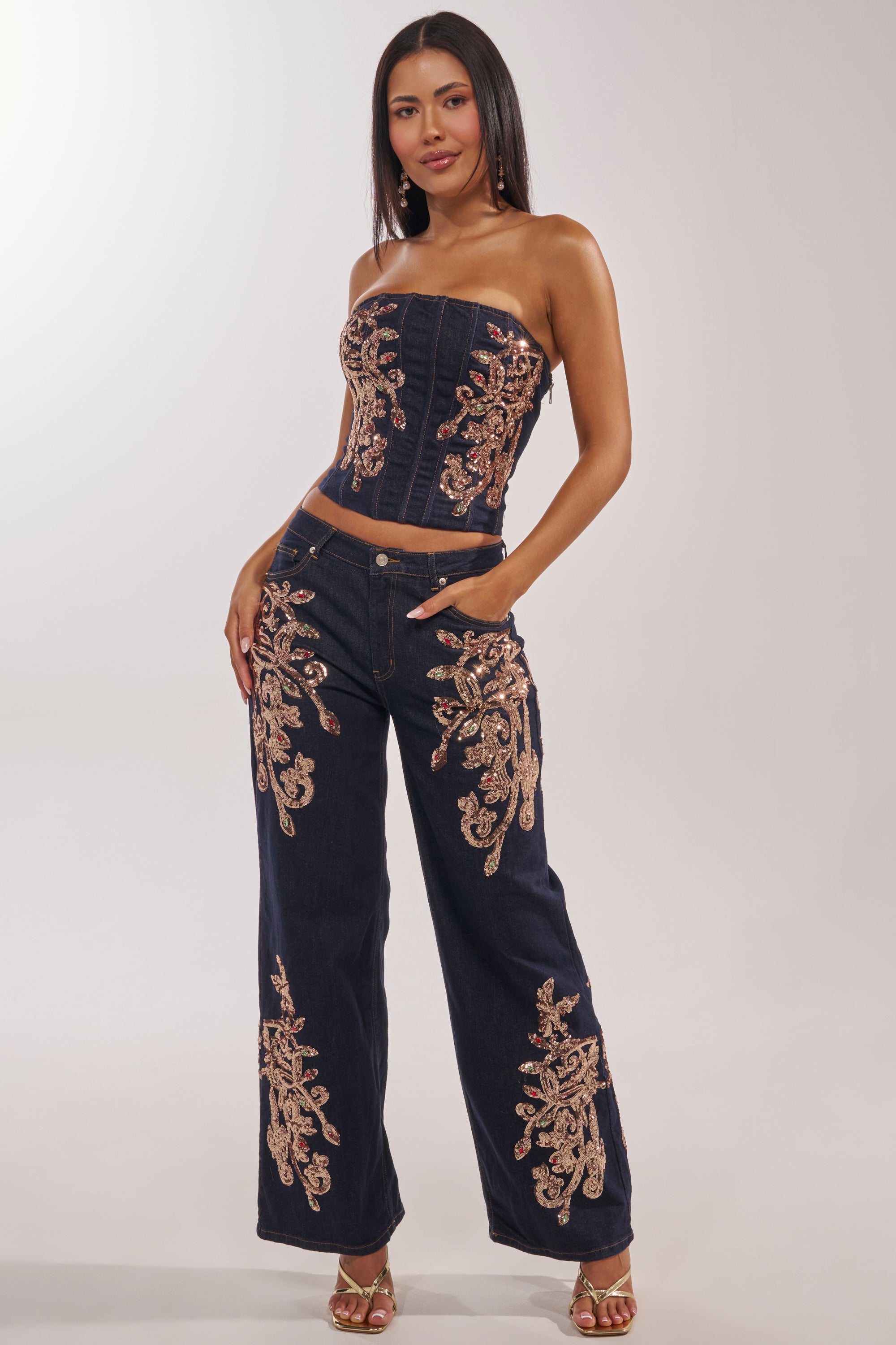 A woman poses in the MALIBU EMBELLISHED WIDE LEG DENIM—a navy blue, wide-leg jean featuring intricate gold embroidery—paired with a matching strapless top and high-heeled sandals, her long dark hair straight in a simple studio setting. 7