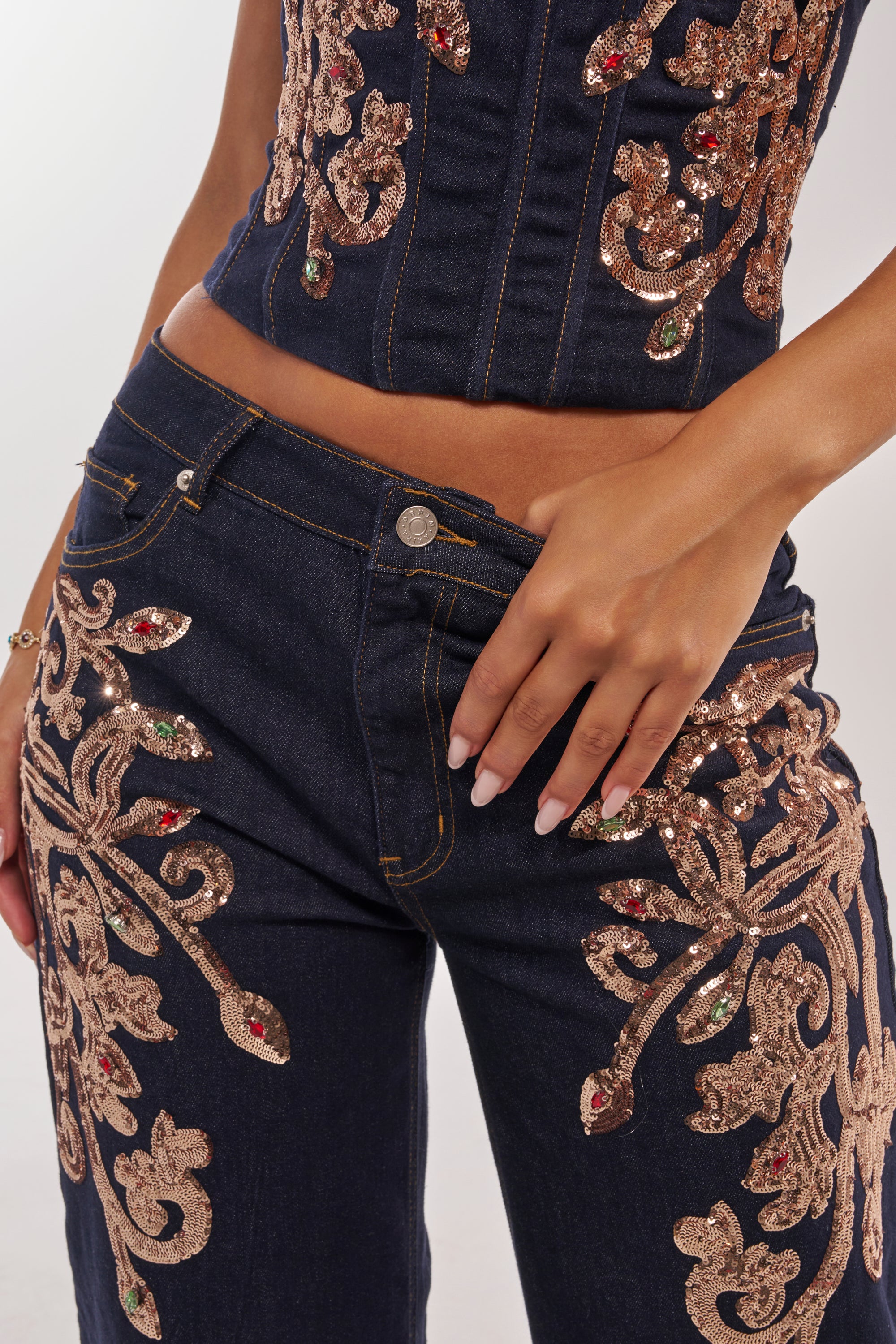 A person wears the MALIBU EMBELLISHED WIDE LEG DENIM and a matching sequin-embroidered corset top, both featuring intricate gold, red, and green floral patterns. Their hand rests on the waistband of the embellished jeans. 8