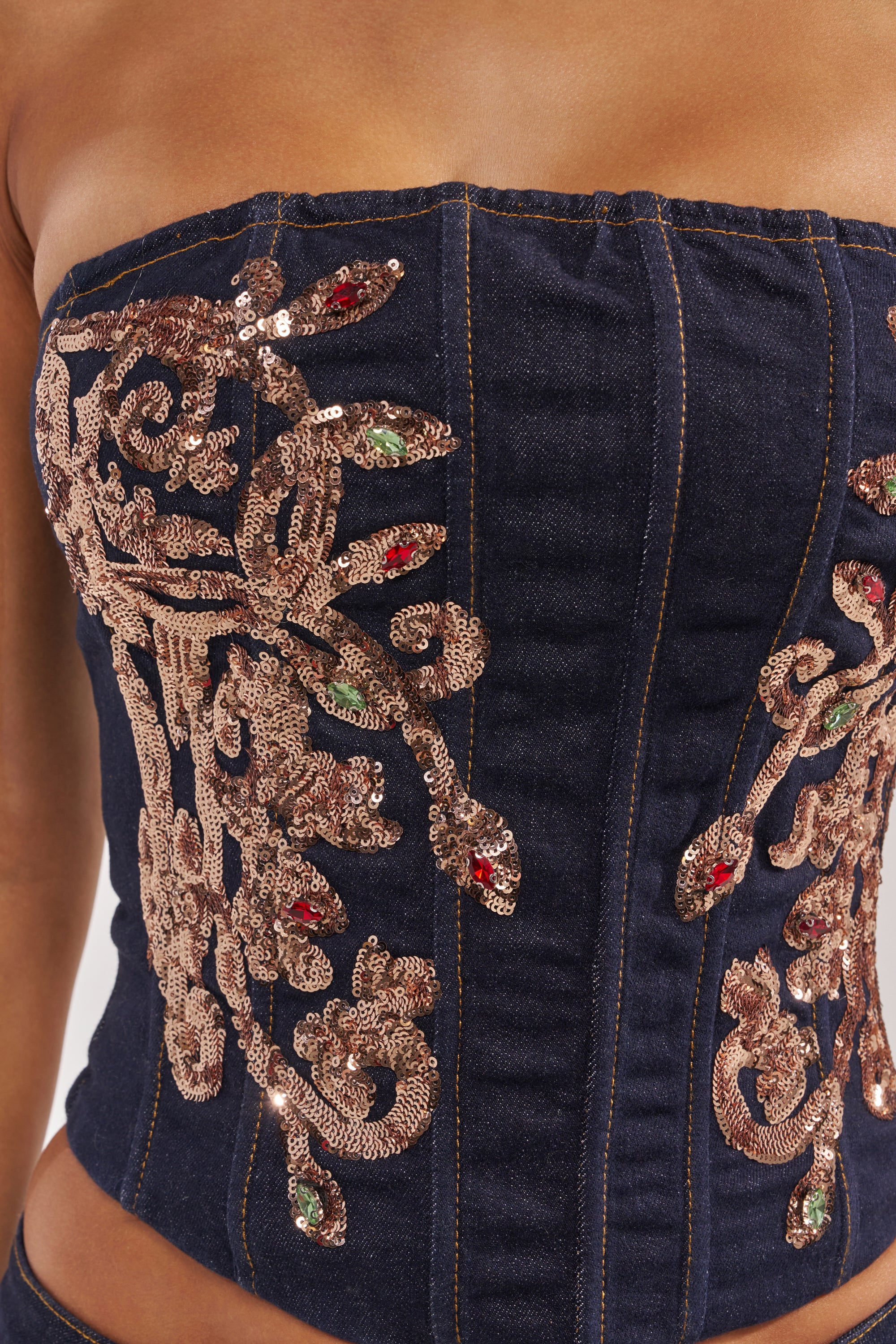 A close-up of a person wearing the MALIBU LACE UP EMBELLISHED DENIM CORSET, strapless and dark blue, featuring ornate gold sequins with green and red bead accents. 9
