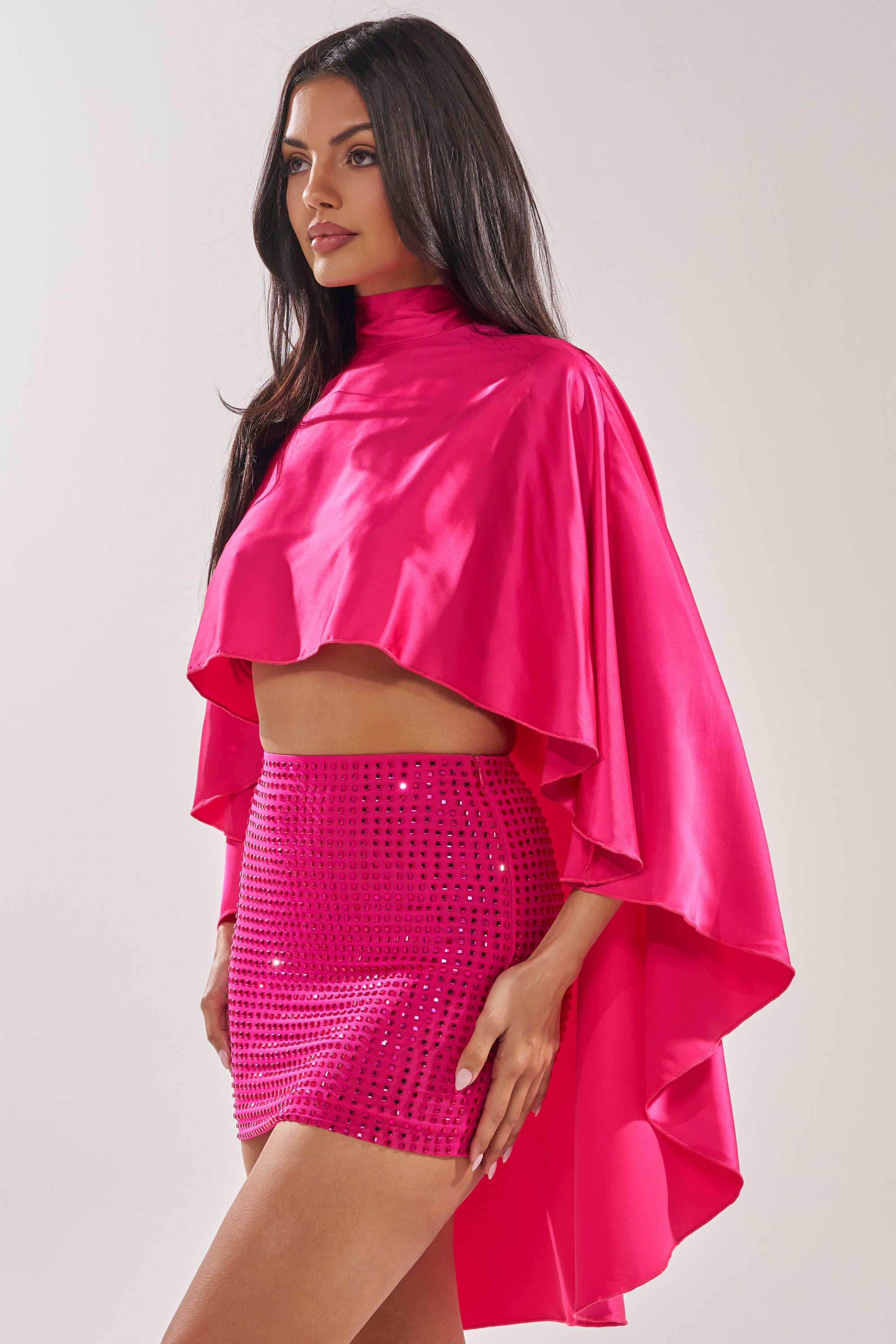 A woman wears the MAKE A MOVE BLOUSE IN FUCHSIA, a vibrant high neck satin cape-style top, paired with a matching sequined mini skirt against a light background. 3
