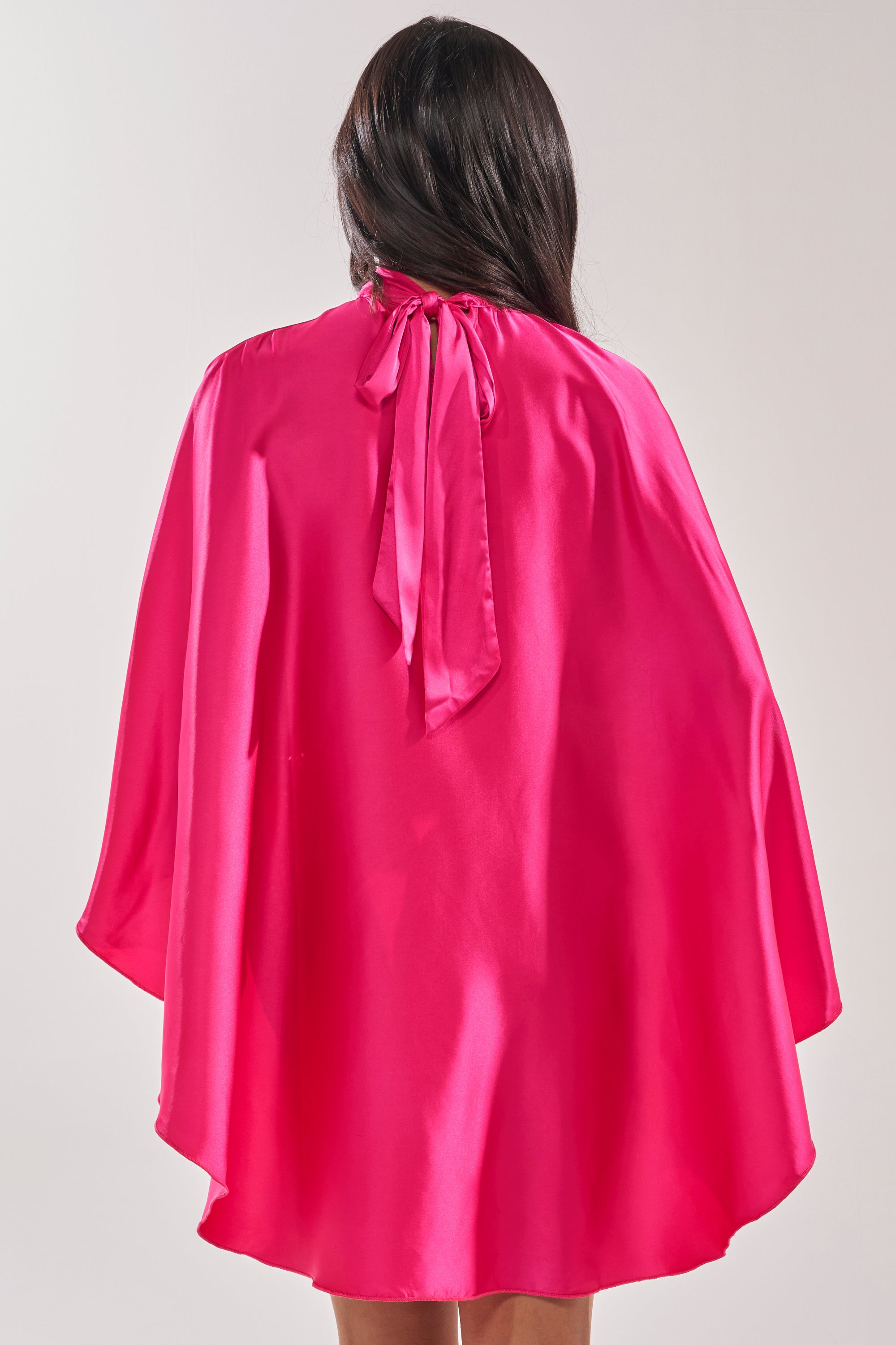 A woman with long dark hair is seen from behind wearing the MAKE A MOVE BLOUSE IN FUCHSIA, a shiny high neck satin blouse styled as a cape with a bow at the neck, draping loosely over her shoulders and back. 4