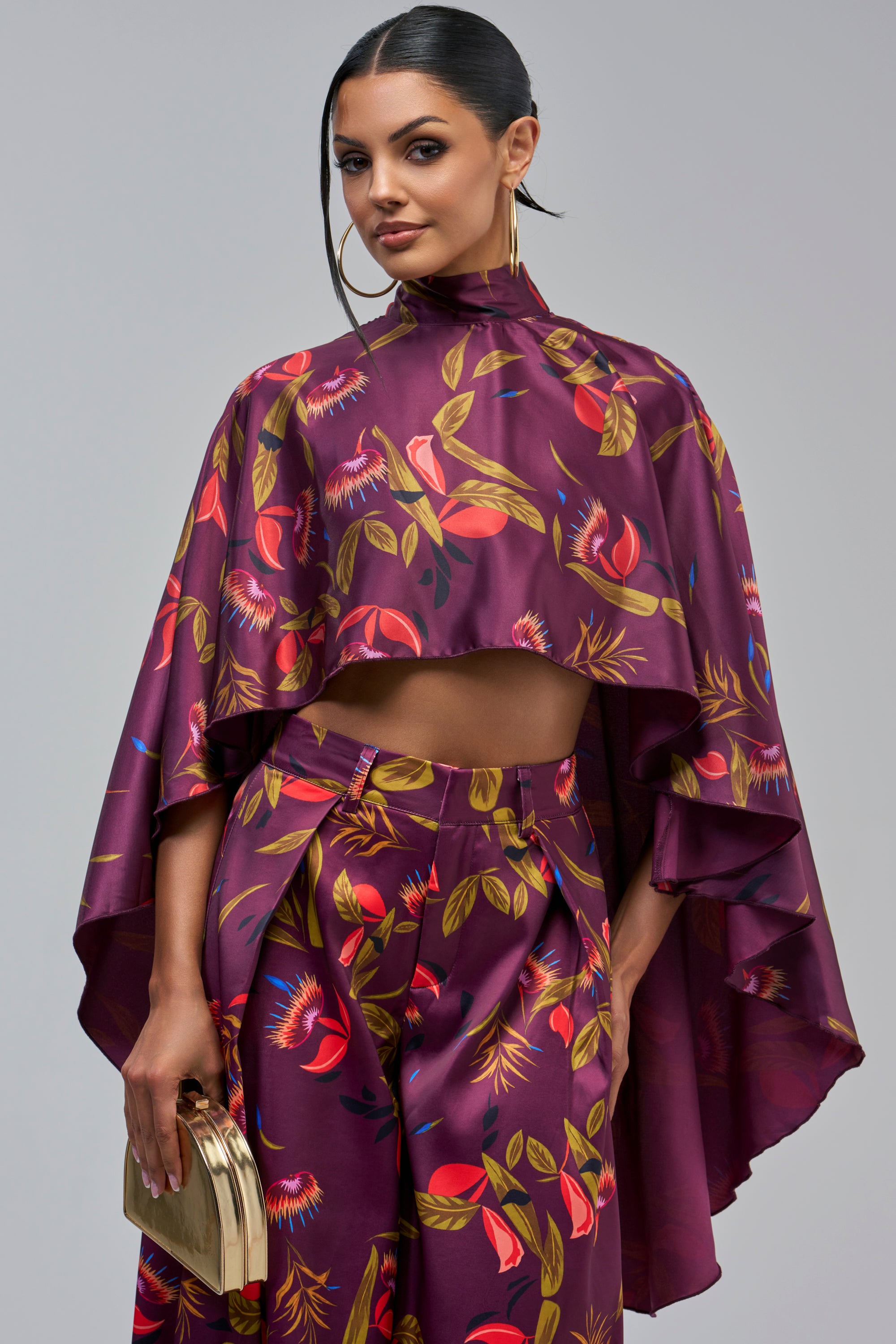A woman wears a PURPLE RAIN SATIN BLOUSE in a purple floral high-neck cape-style cropped design with matching trousers, holding a gold clutch.