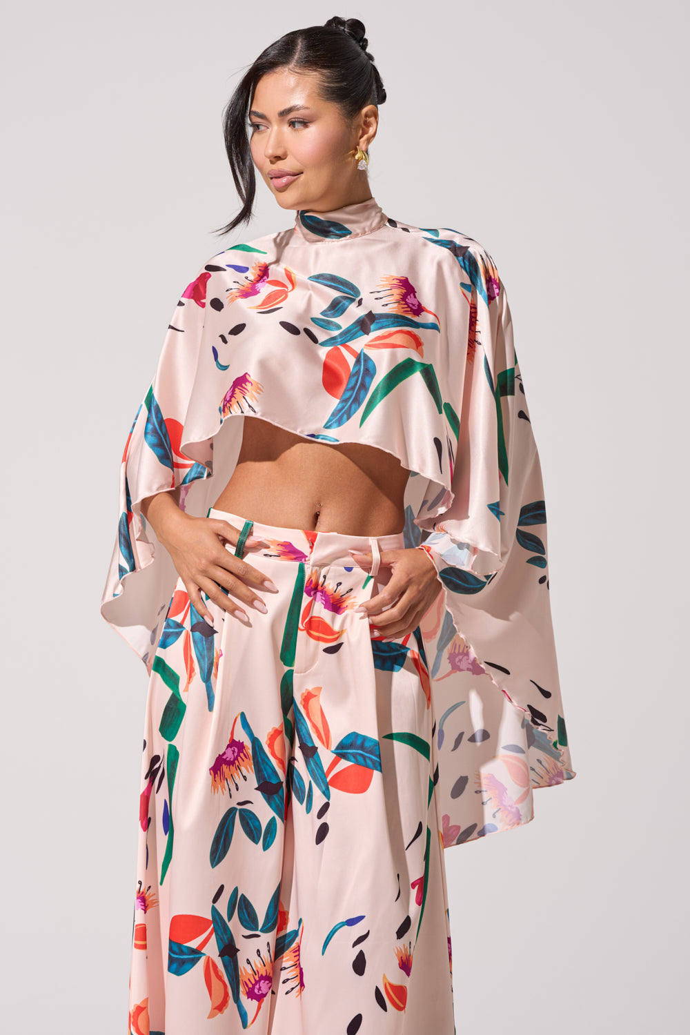 A woman wears the CANDY RAIN SATIN BLOUSE, a shiny, loose-fit floral print top with a cropped cape style, paired with wide-leg pants. She stands against a plain light background, her dark hair styled up and gold earrings completing the look. 0