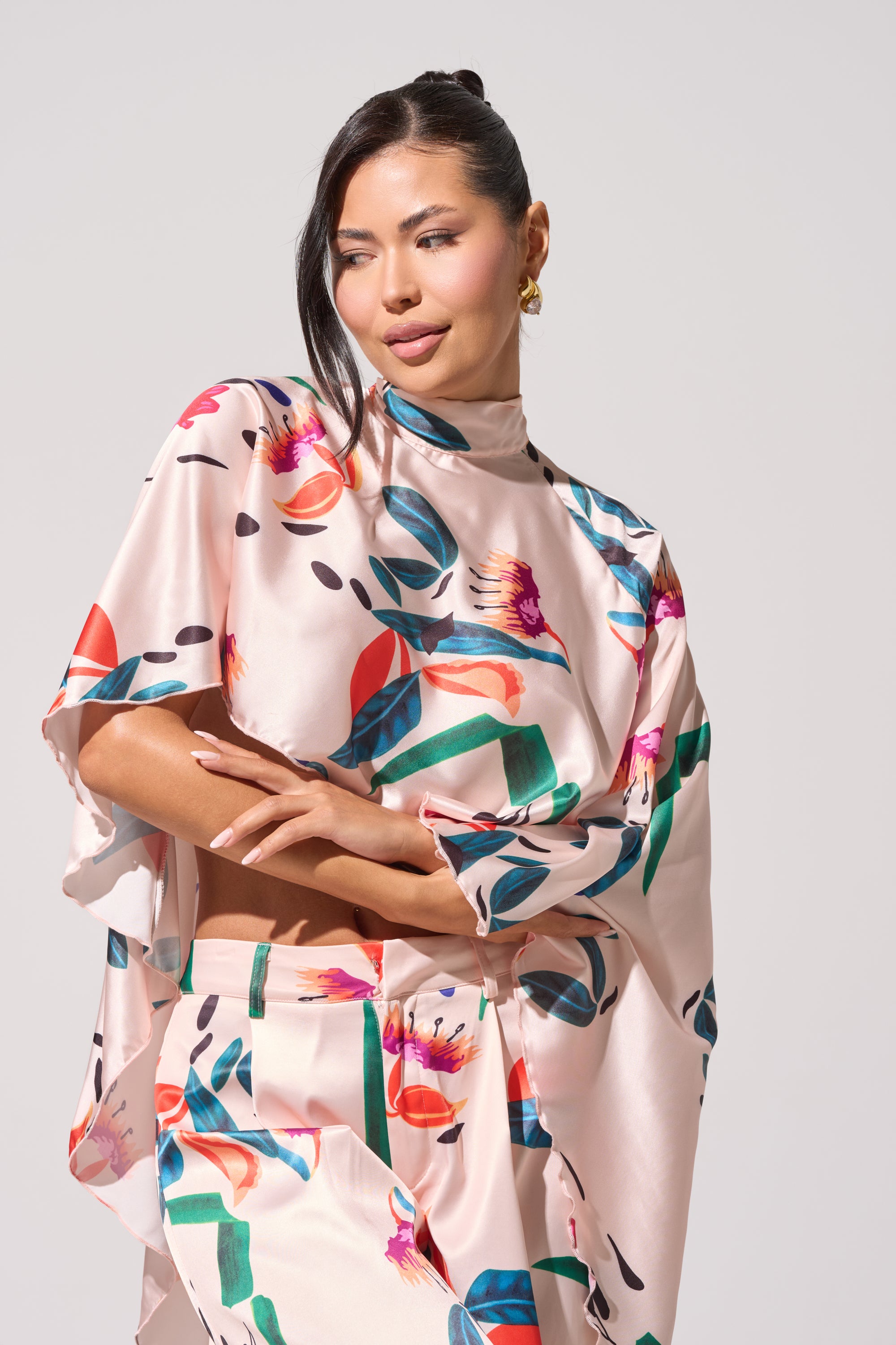 A woman with dark hair in a ponytail and gold earrings wears the CANDY RAIN SATIN BLOUSE, featuring a high-low hem and colorful abstract floral print, posing confidently against a plain light background. 1