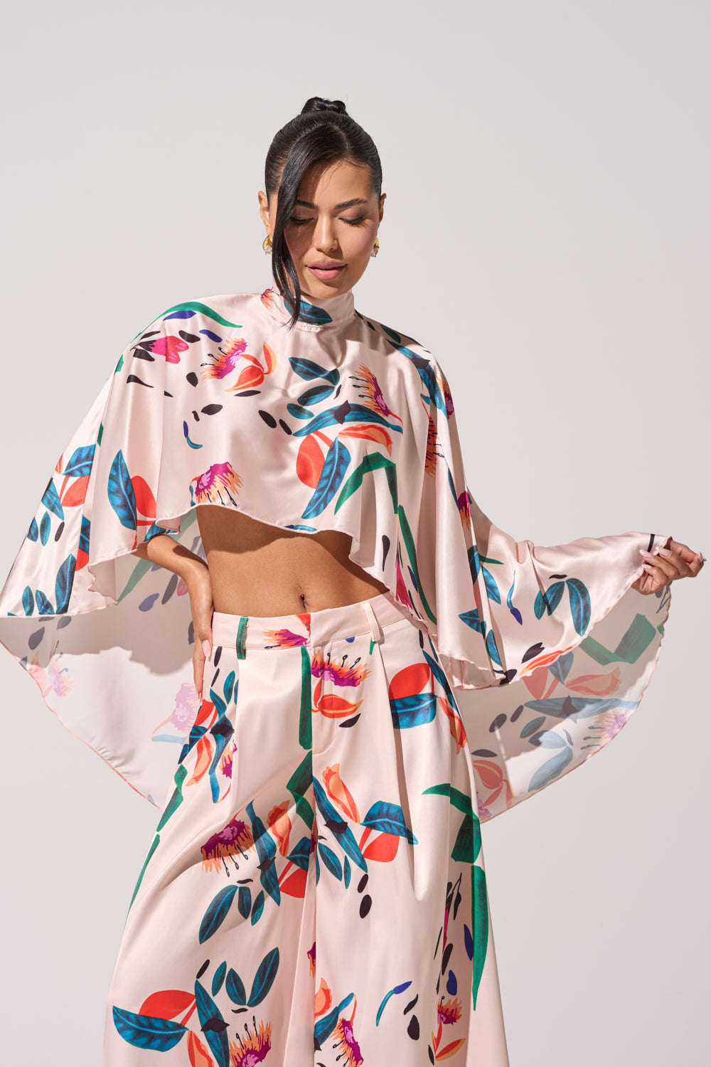 A woman models the CANDY RAIN SATIN BLOUSE, a light pink two-piece with a vibrant floral and leaf print. The cape-style top has loose sleeves and is paired with wide-leg pants. Her hair is styled in a sleek ponytail. 2