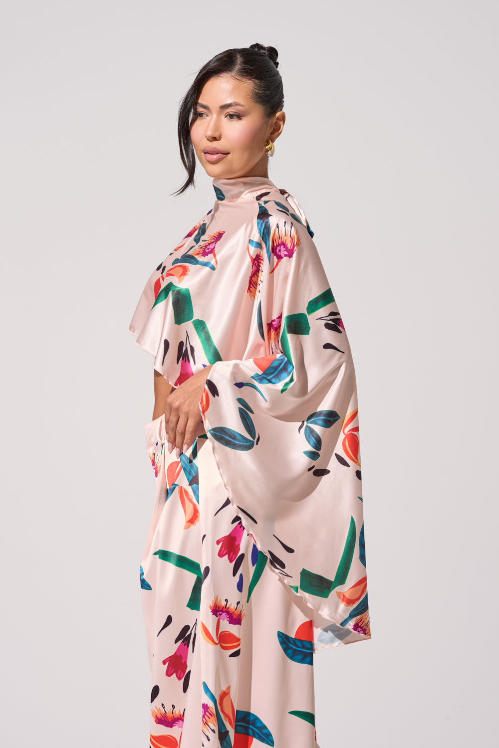 A woman stands in profile, wearing the CANDY RAIN SATIN BLOUSE—a flowing pale pink high-low top with a colorful floral pattern, wide sleeves, and mock neck—her dark hair pulled back against a plain light background. 3