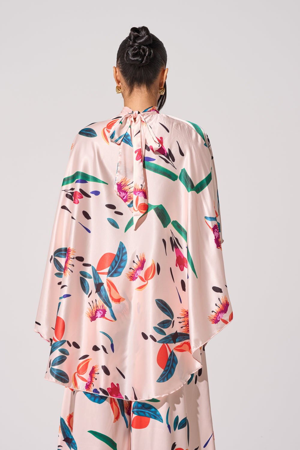 Seen from behind, a person with dark hair in a bun wears the CANDY RAIN SATIN BLOUSE—a loose, light pink high-low top with bold floral and leaf print and a tie-neck. The background is plain light gray. 4