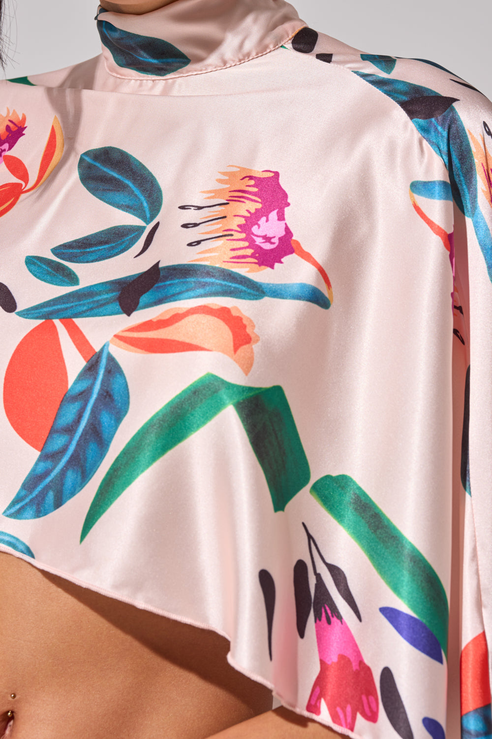 A close-up of a person wearing the CANDY RAIN SATIN BLOUSE, featuring silky light pink satin fabric with a high collar and vibrant green, red, blue, and orange floral prints draping smoothly over the shoulder. 6