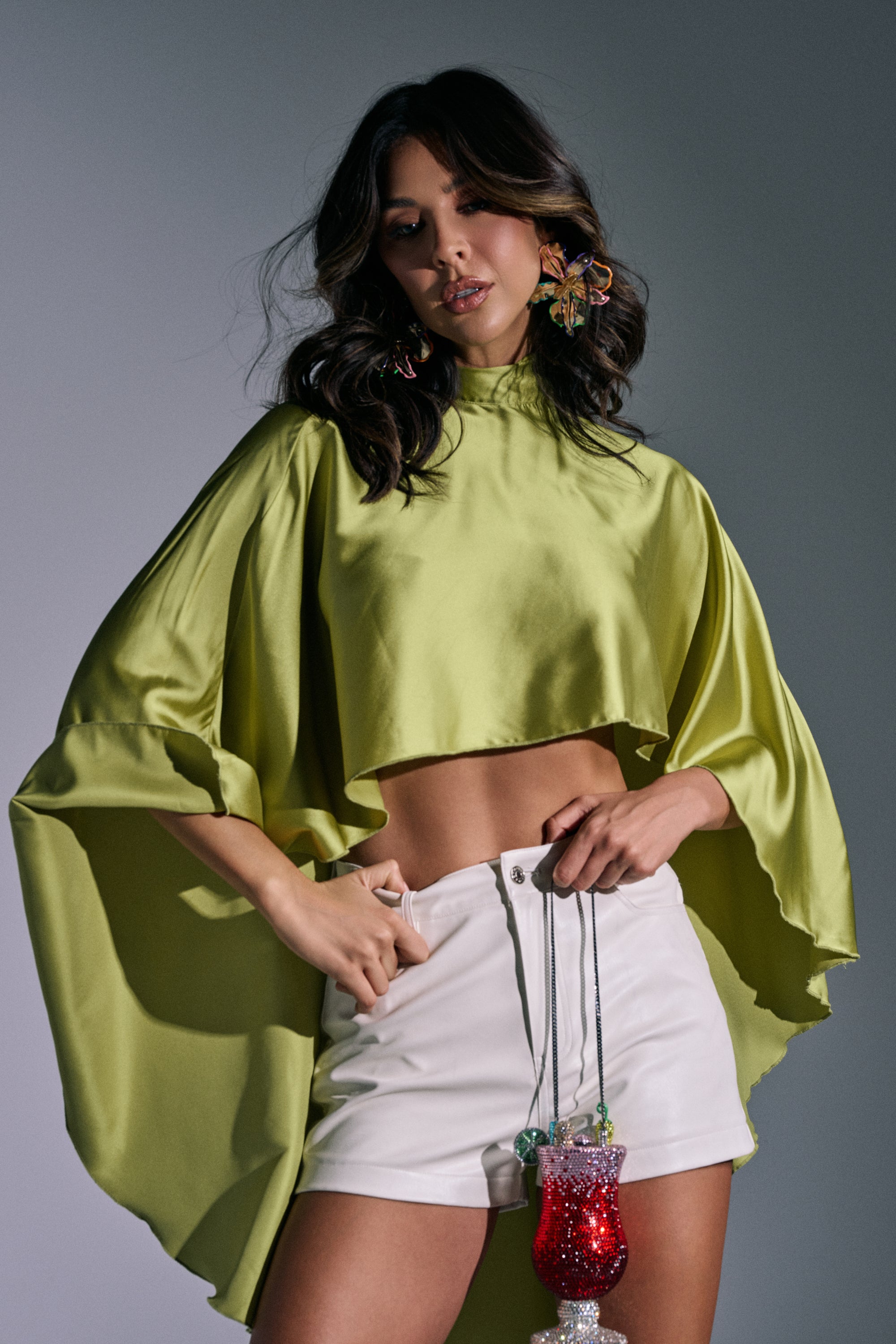 Wearing the BABY MAKE A MOVE BLOUSE IN CHARTREUSE, a woman with wavy dark hair and statement earrings pairs it with high-waisted white shorts, confidently posing while holding two decorative drink cups with straws. 1