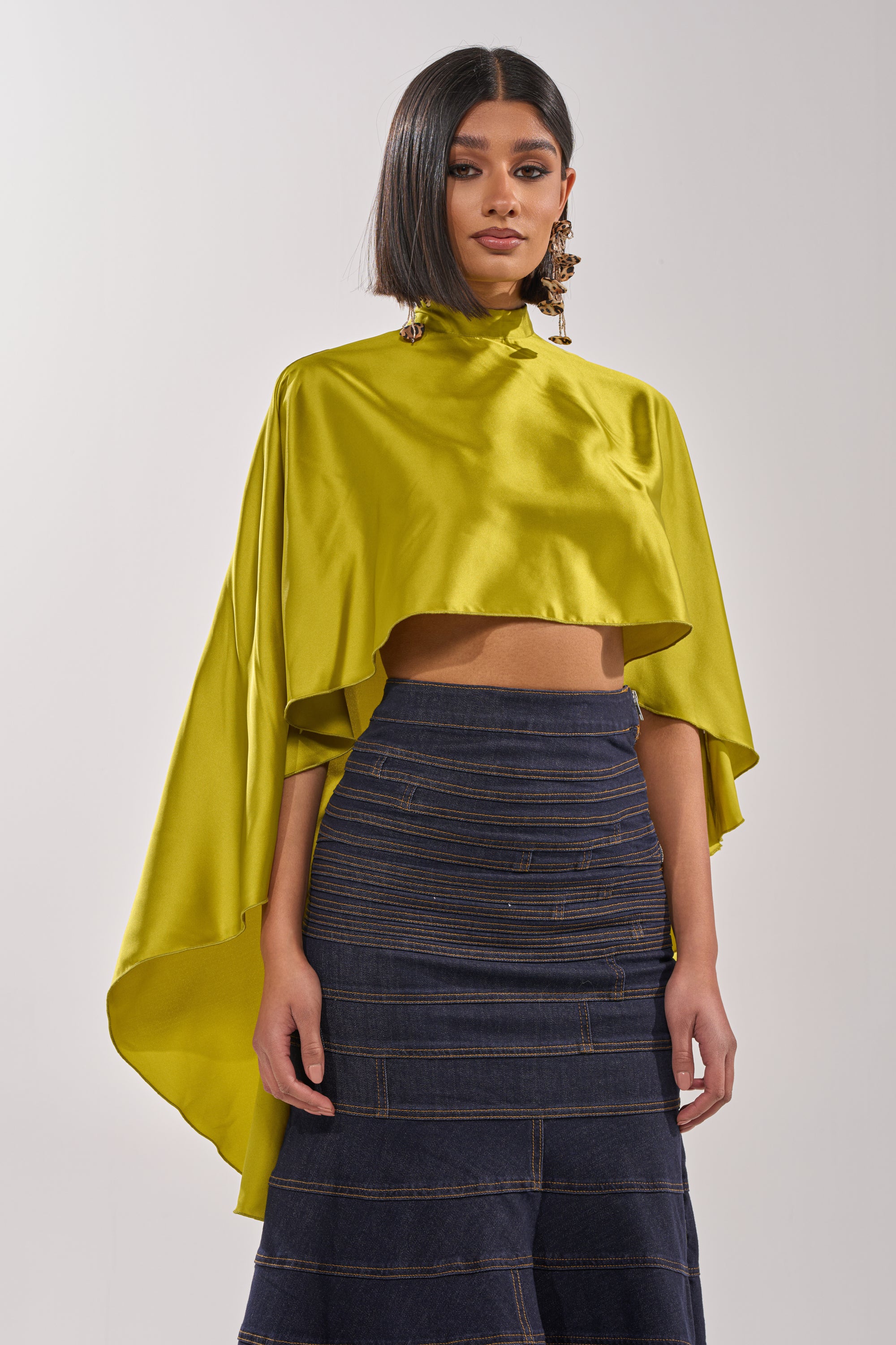 A woman wears the BABY MAKE A MOVE BLOUSE IN CHARTREUSE, a shiny satin mock neck top with cape-style wide sleeves, paired with a dark, fitted tiered skirt. She has straight shoulder-length hair and large statement earrings. 0