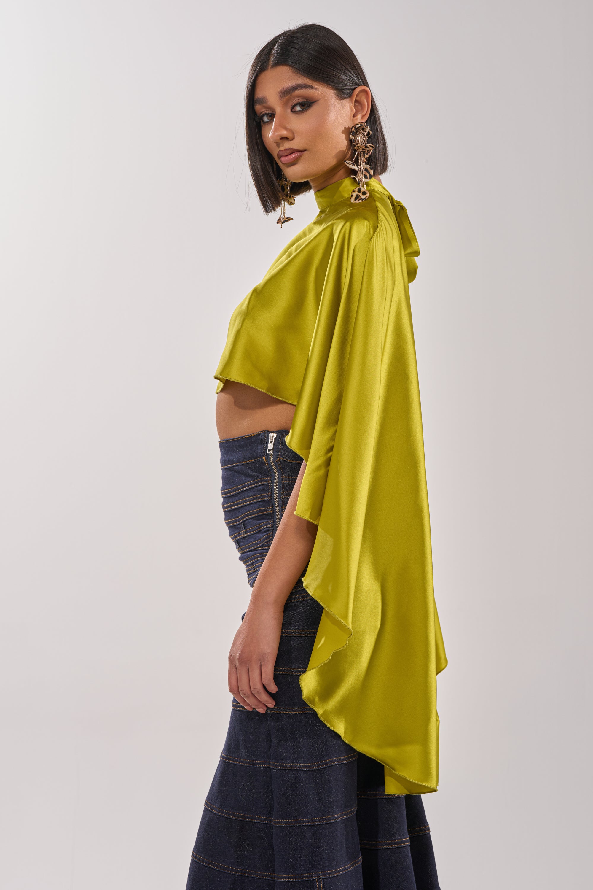 A woman with straight black hair models the BABY MAKE A MOVE BLOUSE IN CHARTREUSE, featuring a shiny satin high neck, draped top, and flowing sleeves. She pairs it with dark flared jeans and statement earrings while posing in profile against a plain background. 6