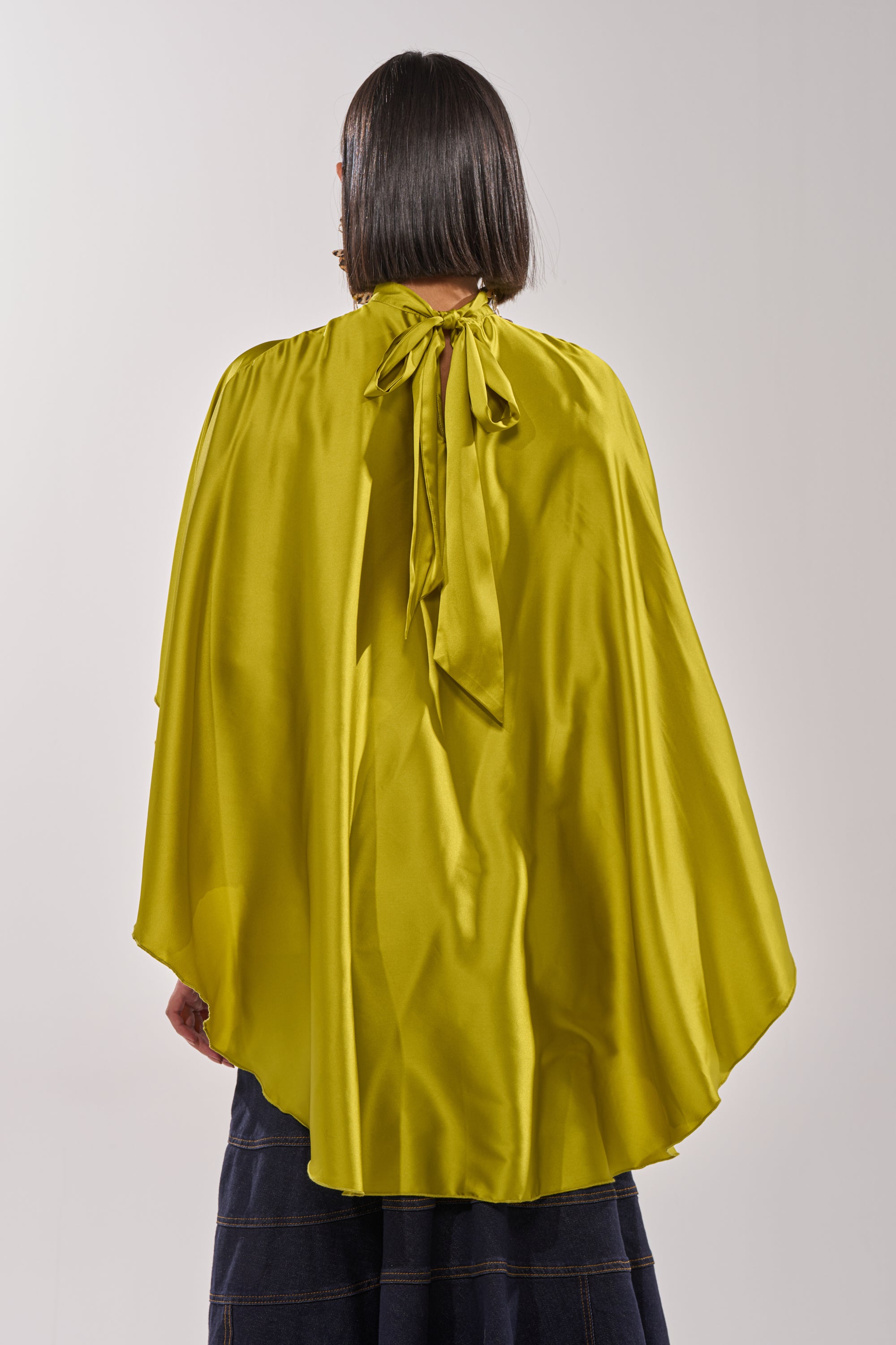 A person with straight, dark, chin-length hair is seen from behind wearing the BABY MAKE A MOVE BLOUSE IN CHARTREUSE, styled as a shiny yellow-green cape with a bow at the neck. The lower part of dark bottoms is visible. 7