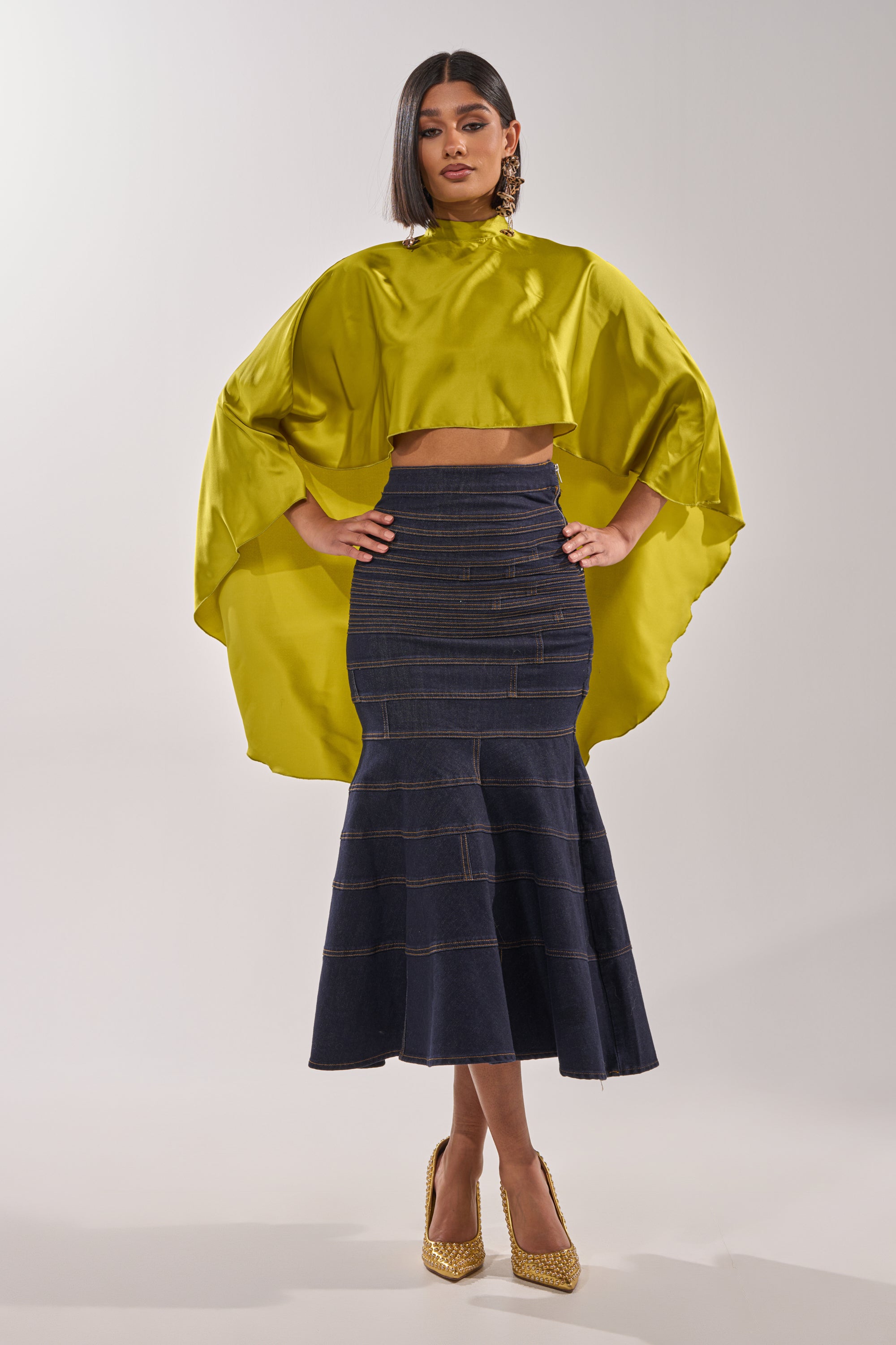 A woman stands confidently in the BABY MAKE A MOVE BLOUSE IN CHARTREUSE, featuring shiny satin fabric and draped sleeves, paired with a high-waisted dark denim mermaid skirt, gold glitter heels, straight dark hair, and statement earrings. 8