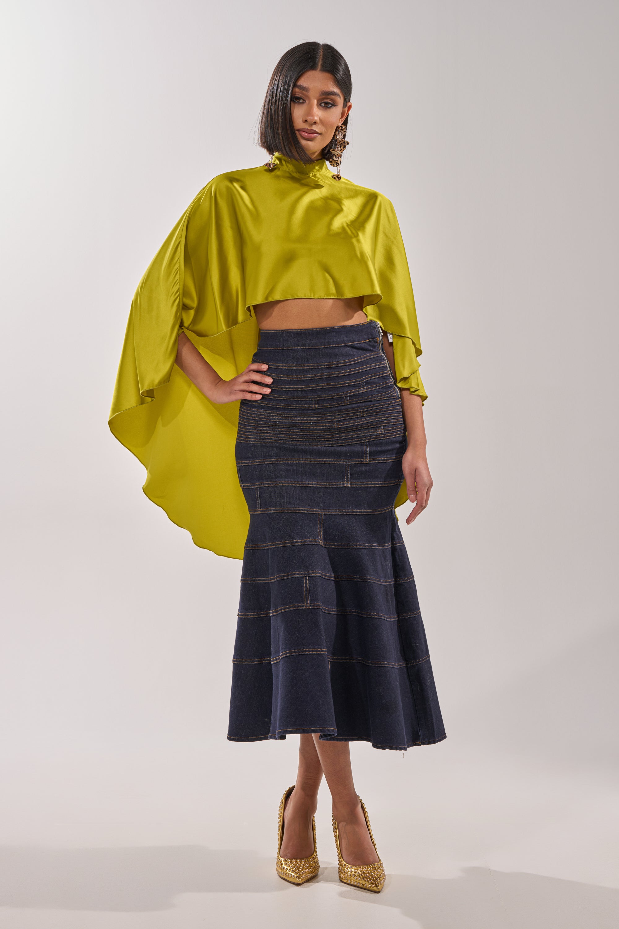 A woman models the BABY MAKE A MOVE BLOUSE IN CHARTREUSE, a satin mock neck top with a cape-style high-low cropped design, paired with a fitted dark blue flared midi skirt, gold heels, and statement earrings against a plain backdrop. 9