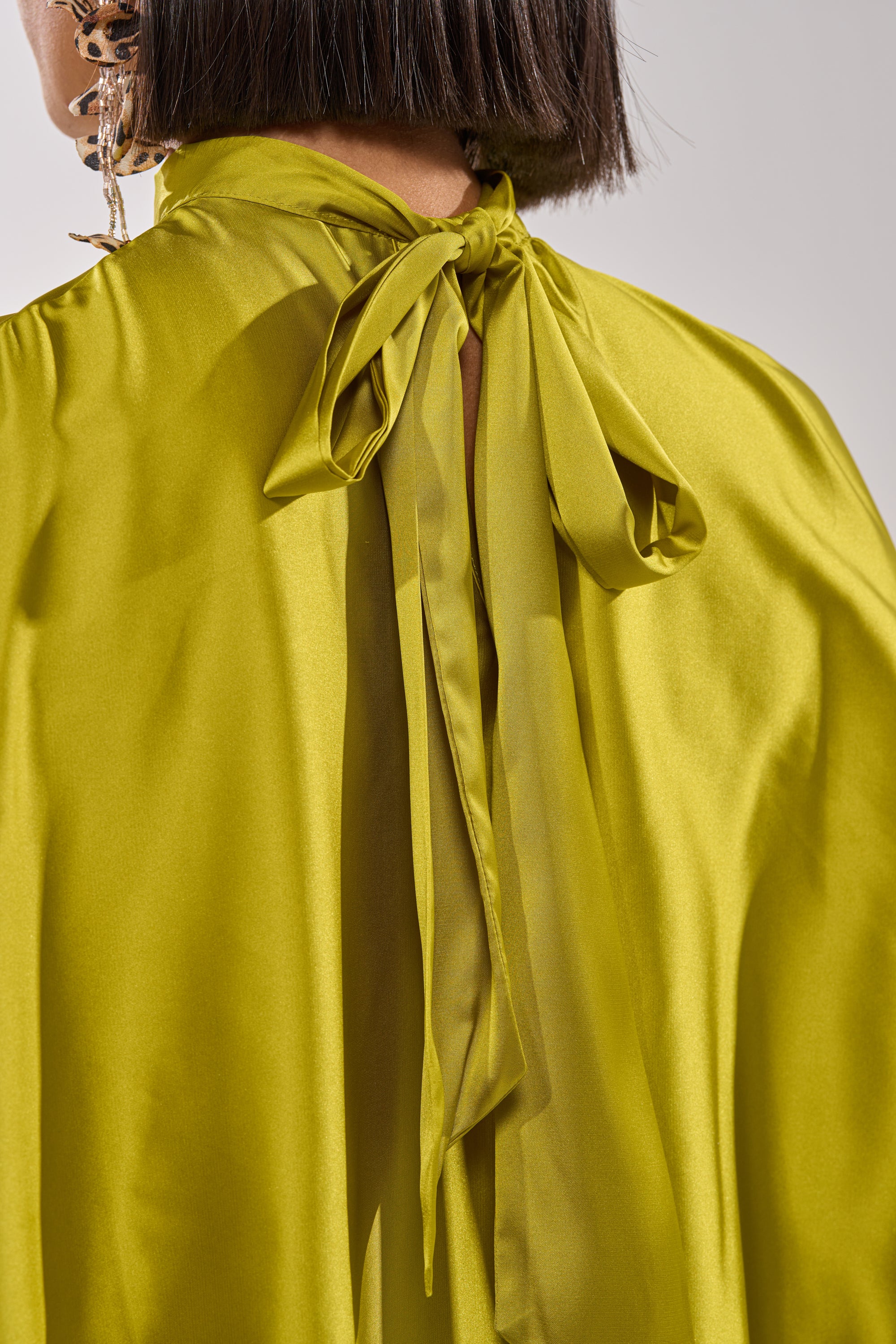 Close-up of a person wearing the BABY MAKE A MOVE BLOUSE IN CHARTREUSE, featuring shiny satin fabric and a bow tied at the back of the mock neck. Visible are their short, dark hair and part of a dangling earring. 11
