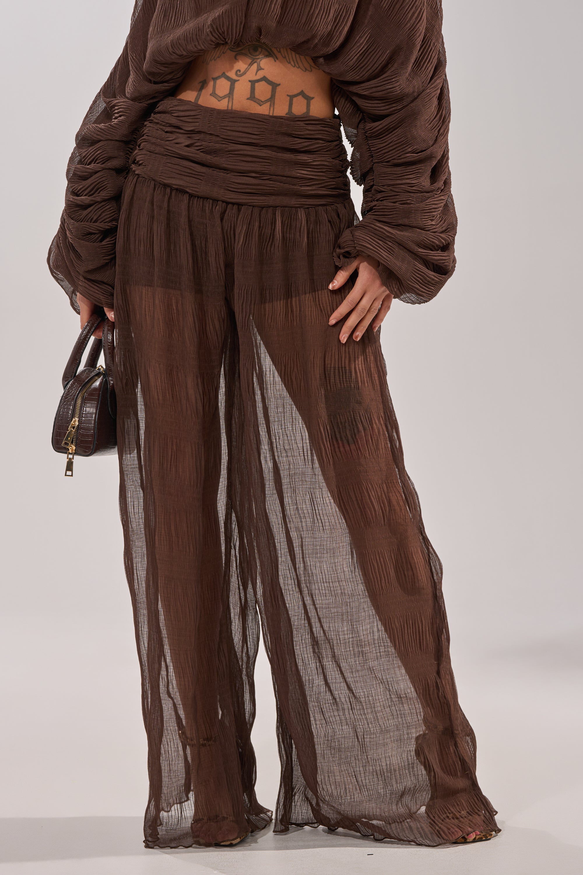 Wearing the BABY THATS HOW I WANT IT PALAZZO PANT in brown, paired with a matching textured top, a person holds a small brown handbag. The loose, sheer layered pants feature a high-rise smocked waistband and reveal their tattooed midriff. 3