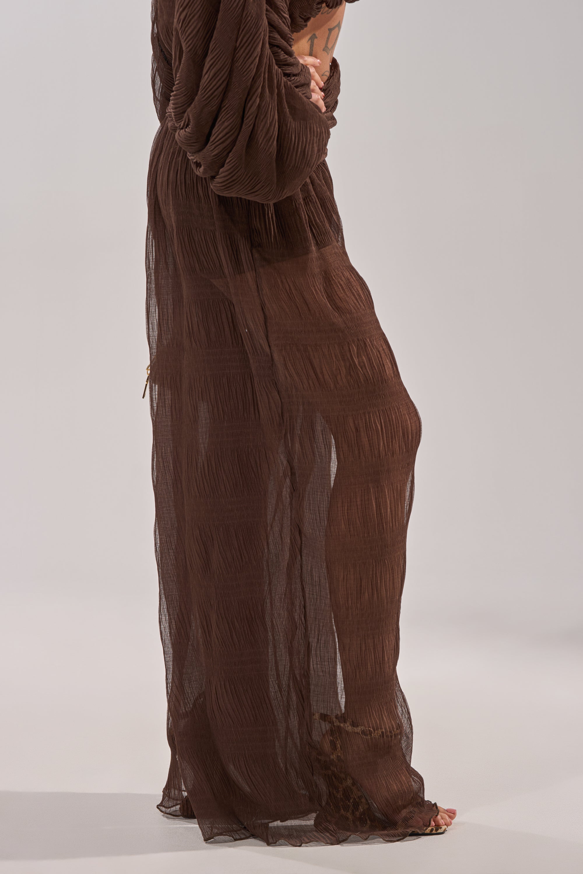 A person stands barefoot in the BABY THATS HOW I WANT IT PALAZZO PANT, featuring a high-rise smocked waistband and sheer, textured brown layers that drape loosely over their lower half against a neutral background. 5