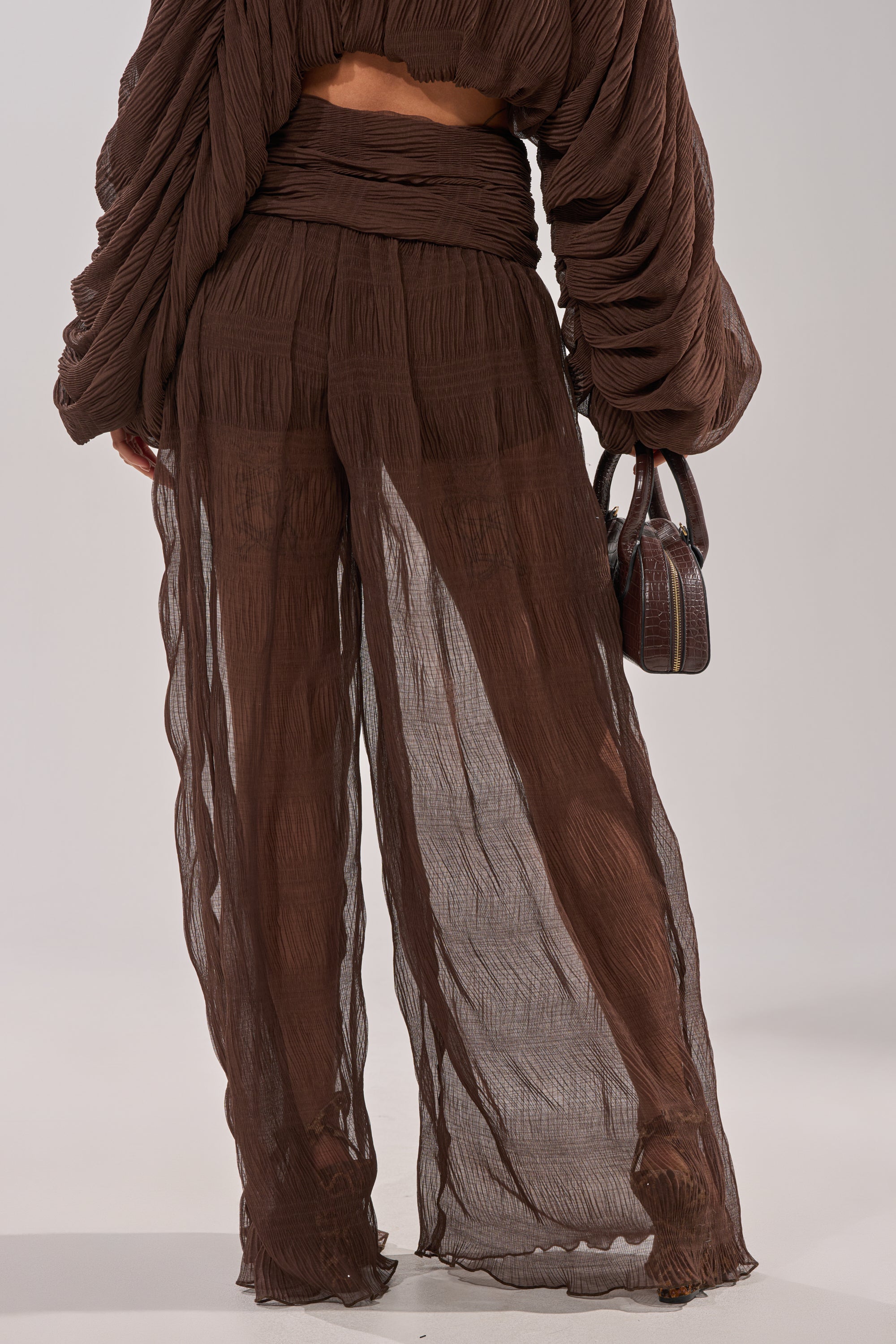 Wearing the BABY THATS HOW I WANT IT PALAZZO PANT—sheer, layered brown high-rise pants with a smocked waistband—matched with a long-sleeved top and holding a small brown handbag. The flowy, semi-transparent look is viewed from the back. 6