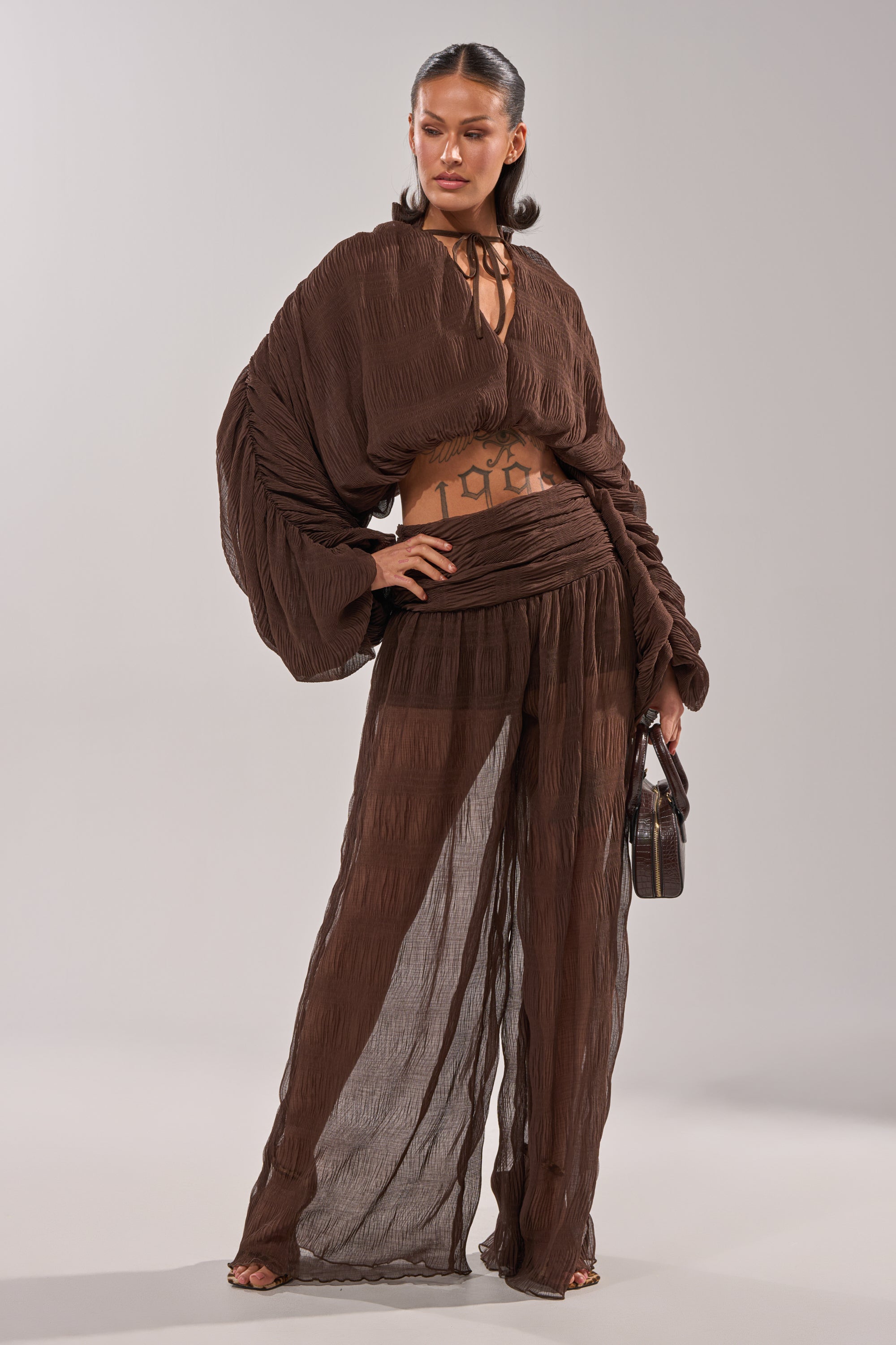 A woman confidently models the BABY THATS HOW I WANT IT PALAZZO PANT, a sheer, textured brown two-piece with wide-leg pants and a loose long-sleeved top. She holds a small handbag, her hair slicked back, revealing her midriff and tattoos. 7