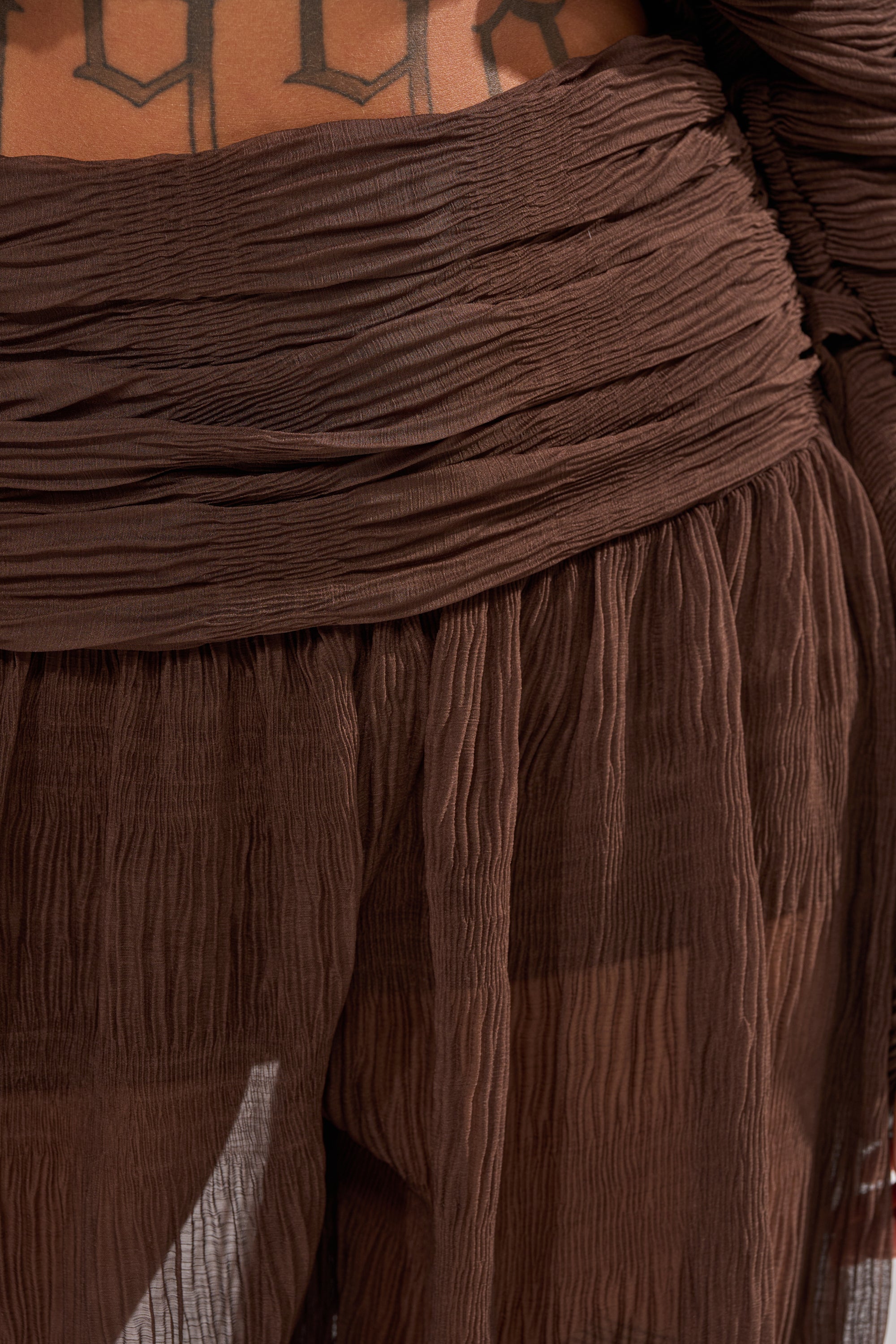 A close-up of a person's midsection wearing the BABY THATS HOW I WANT IT PALAZZO PANT, featuring brown textured sheer layers and a high-rise smocked waistband. Black tattooed letters show above the fabric on the lower torso. 8