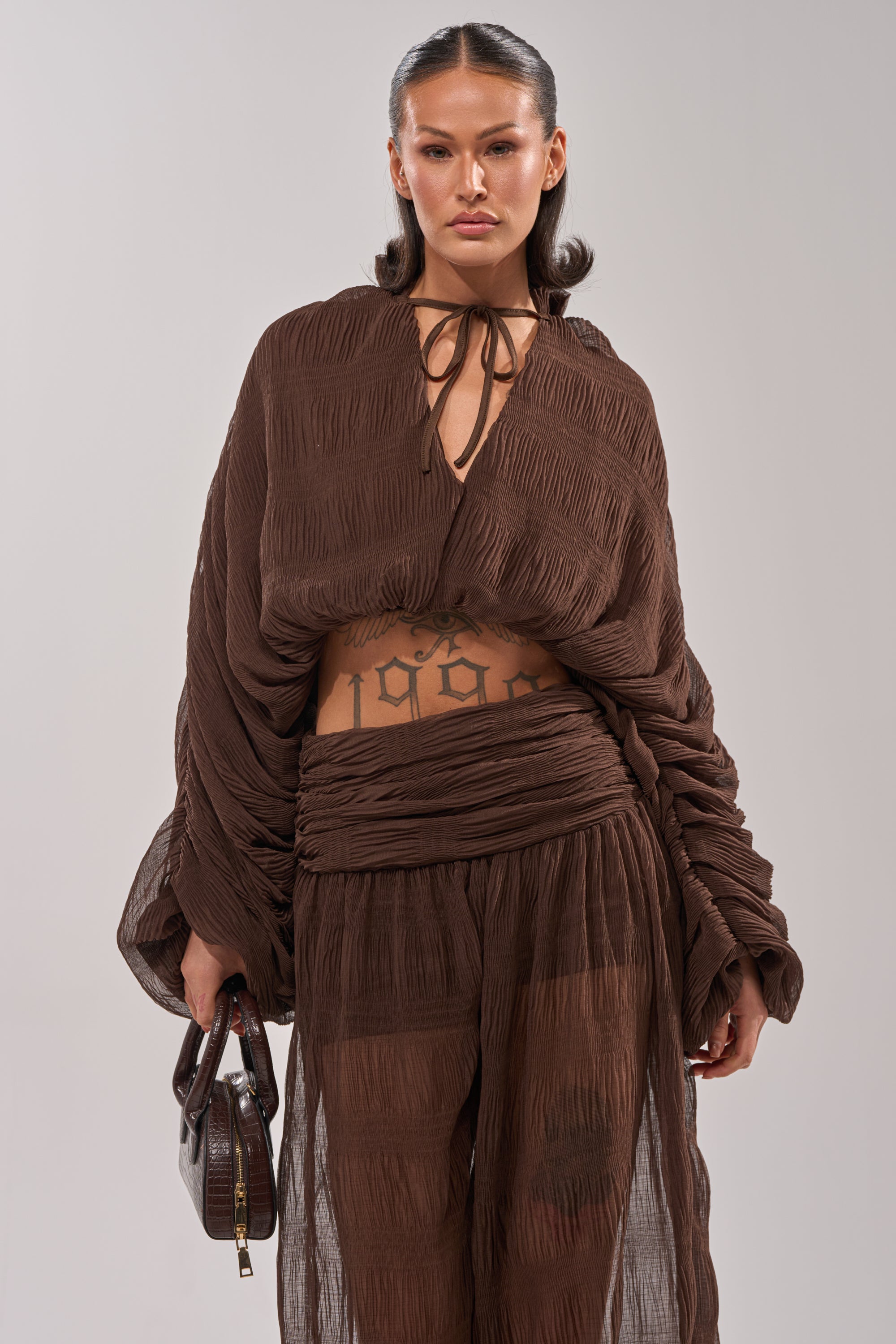 A woman stands confidently in the BABY THATS HOW I WANT IT BLOUSE, a sheer brown balloon-style top with wide sleeves, paired with matching pants and a small brown handbag. Her neatly styled hair and a visible torso tattoo complete the look. 0