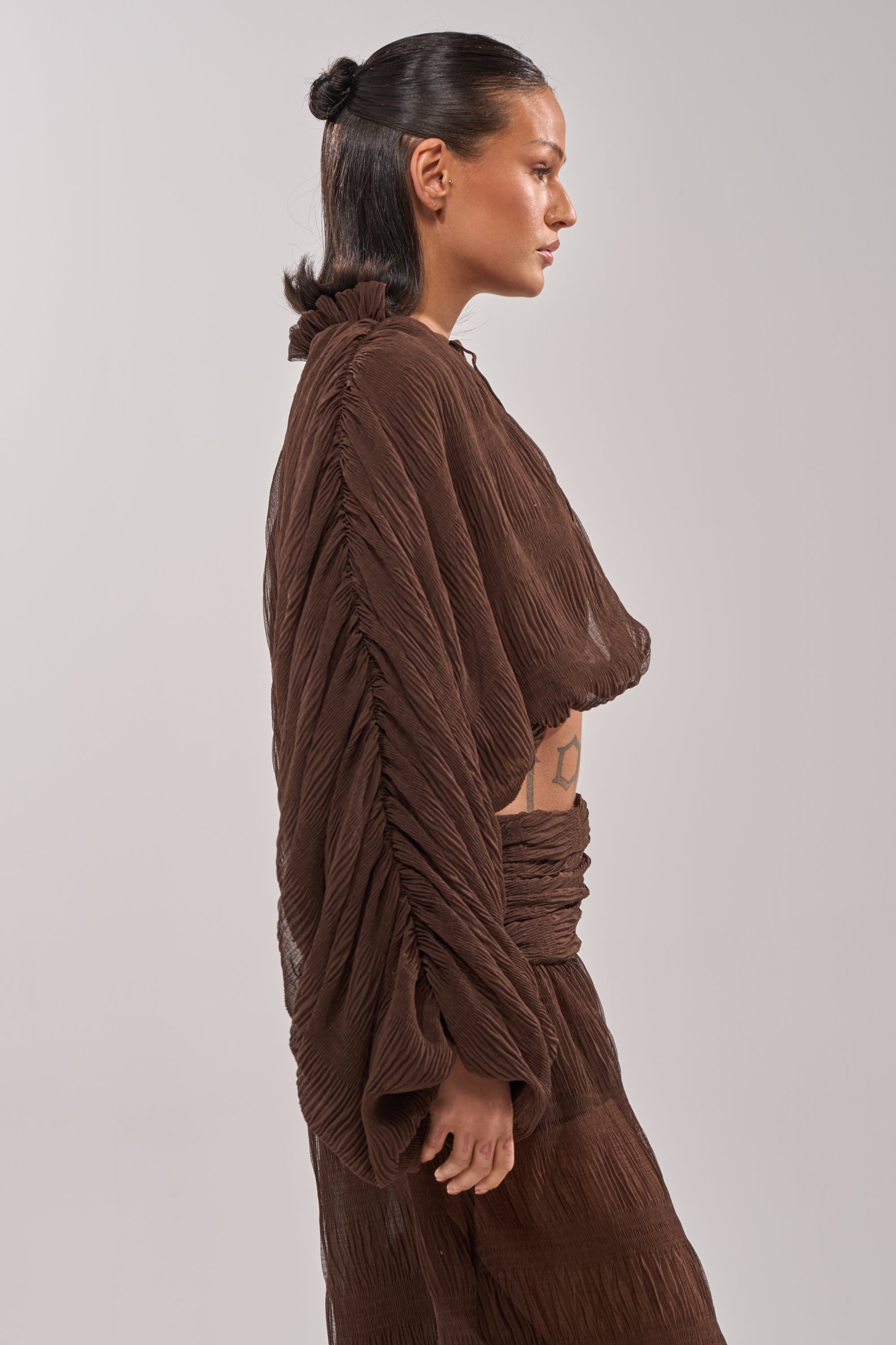 A person with dark hair in a low bun models the BABY THATS HOW I WANT IT BLOUSE and matching skirt in sheer brown, standing in profile against a plain light background. 2
