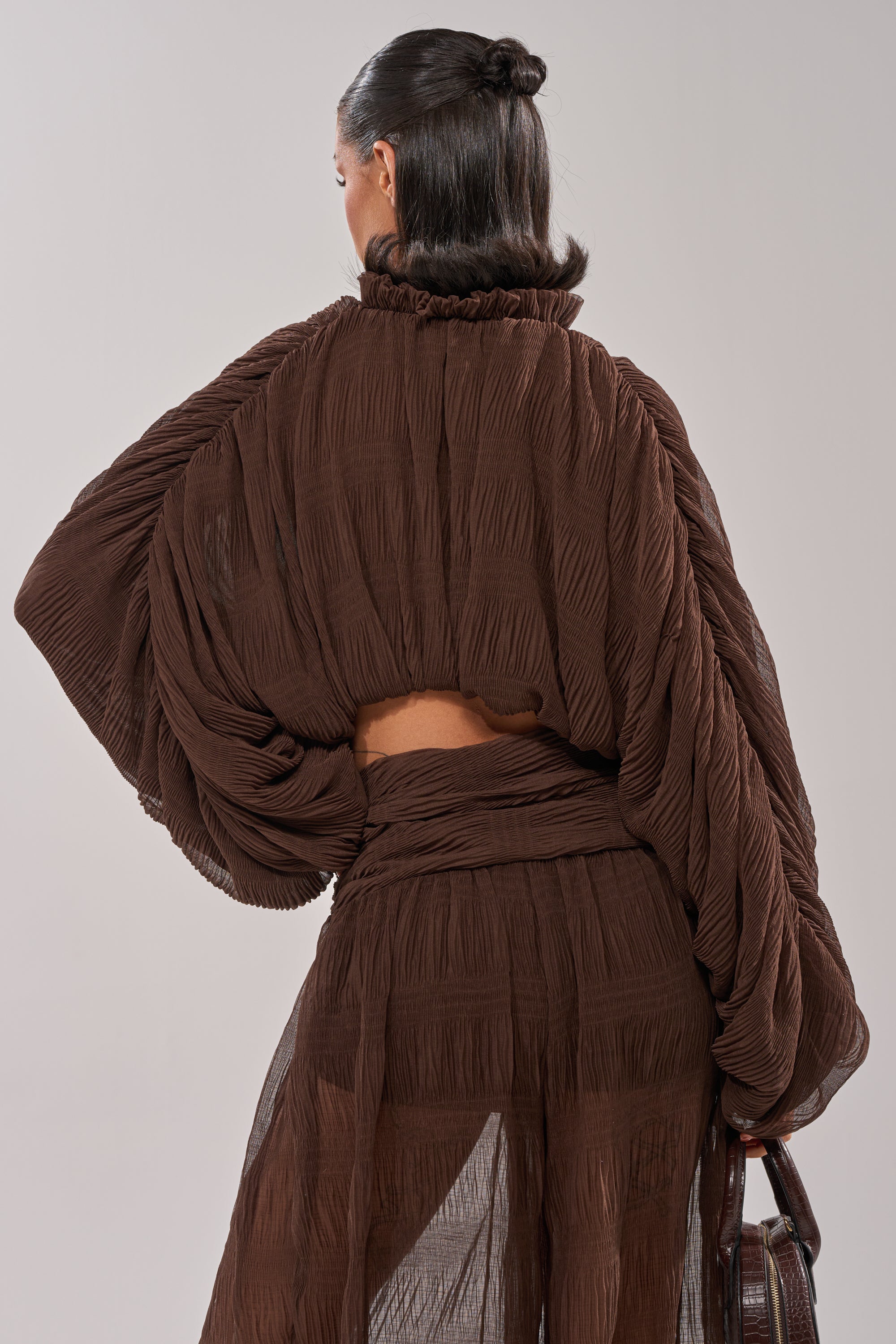 A woman with dark hair in a bun, turned from the camera, wears the sheer brown BABY THATS HOW I WANT IT BLOUSE and matching skirt, holding a dark bag in her left hand against a light background. 3