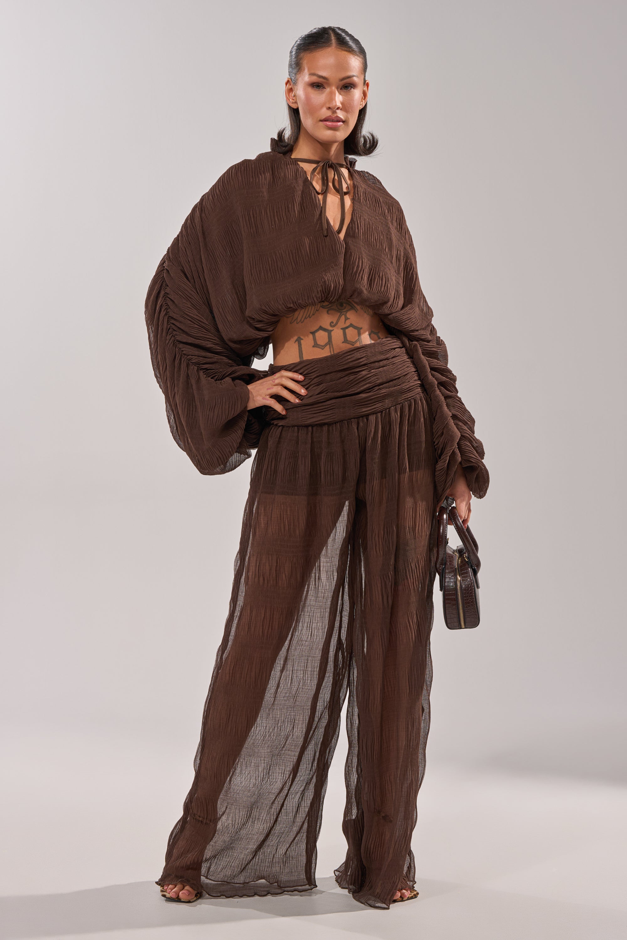A woman stands confidently in the BABY THATS HOW I WANT IT BLOUSE, a sheer brown balloon blouse with a billowy cropped silhouette. She holds a small brown handbag and her slicked-back hair reveals tattoos on her midriff. 4